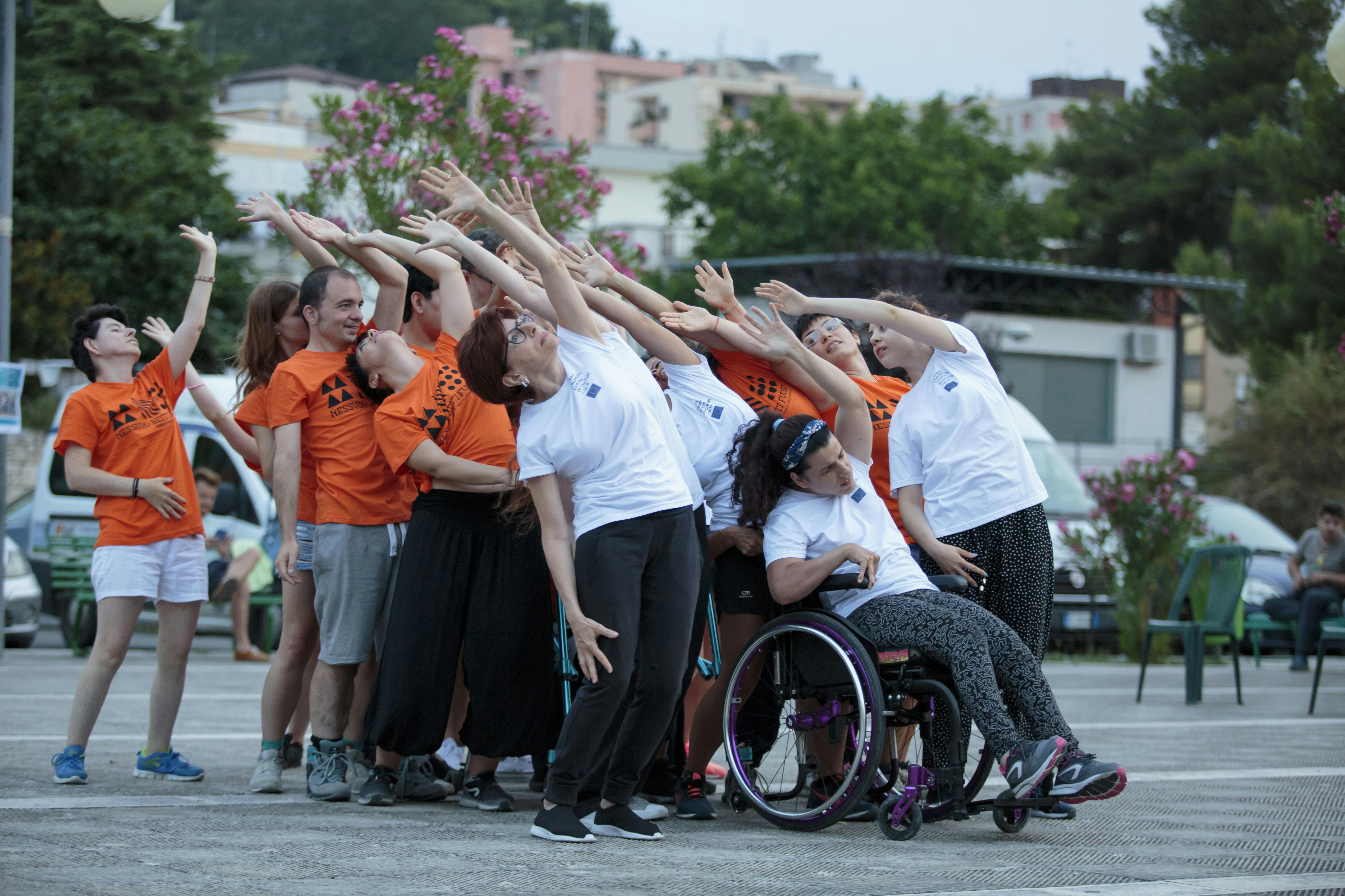 A group of performers with and without disabilities during a performance. They raise one arm upward while slightly tilting their torso back.