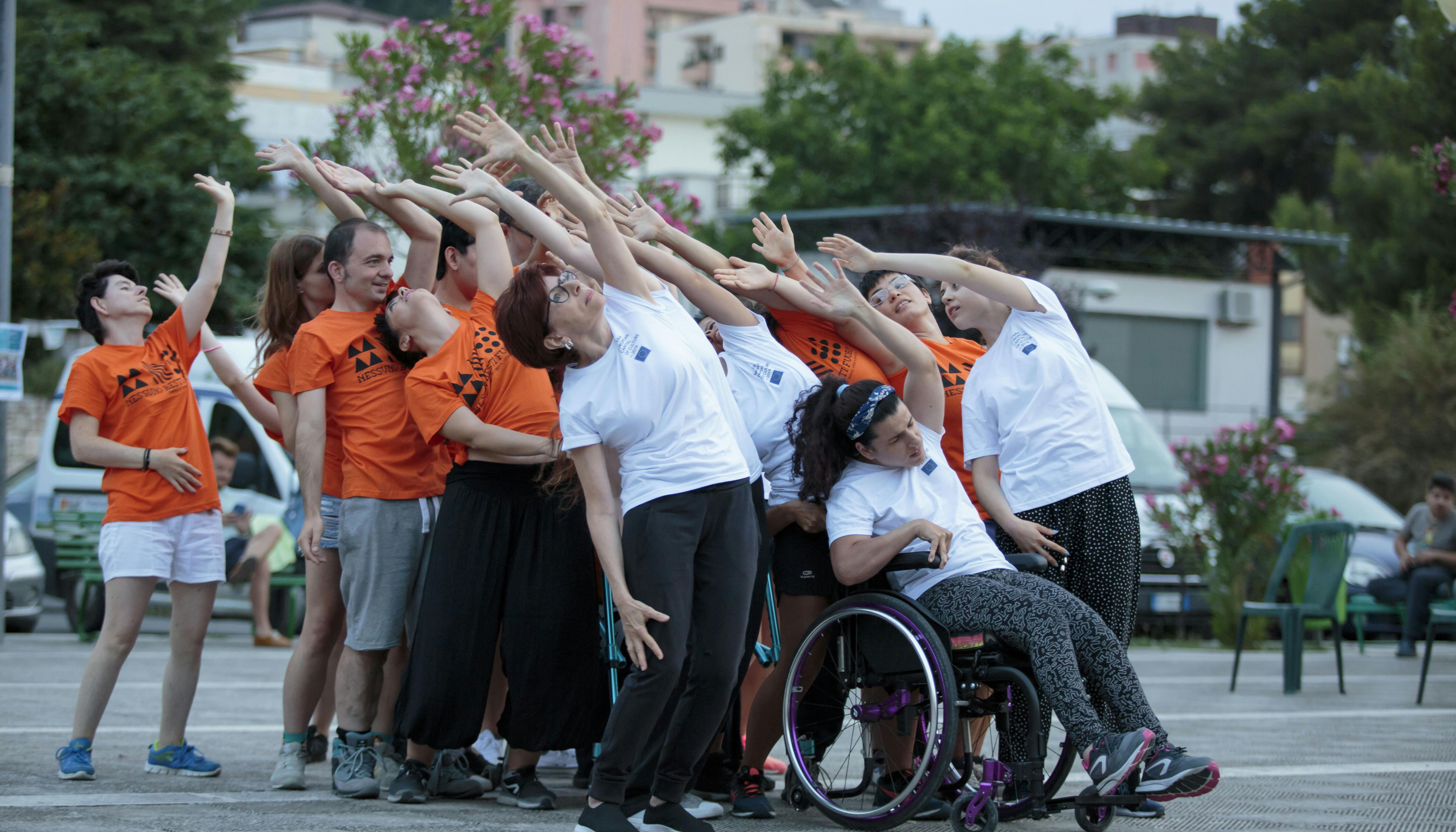 A group of performers with and without disabilities during a performance. They raise one arm upward while slightly tilting their torso back.
