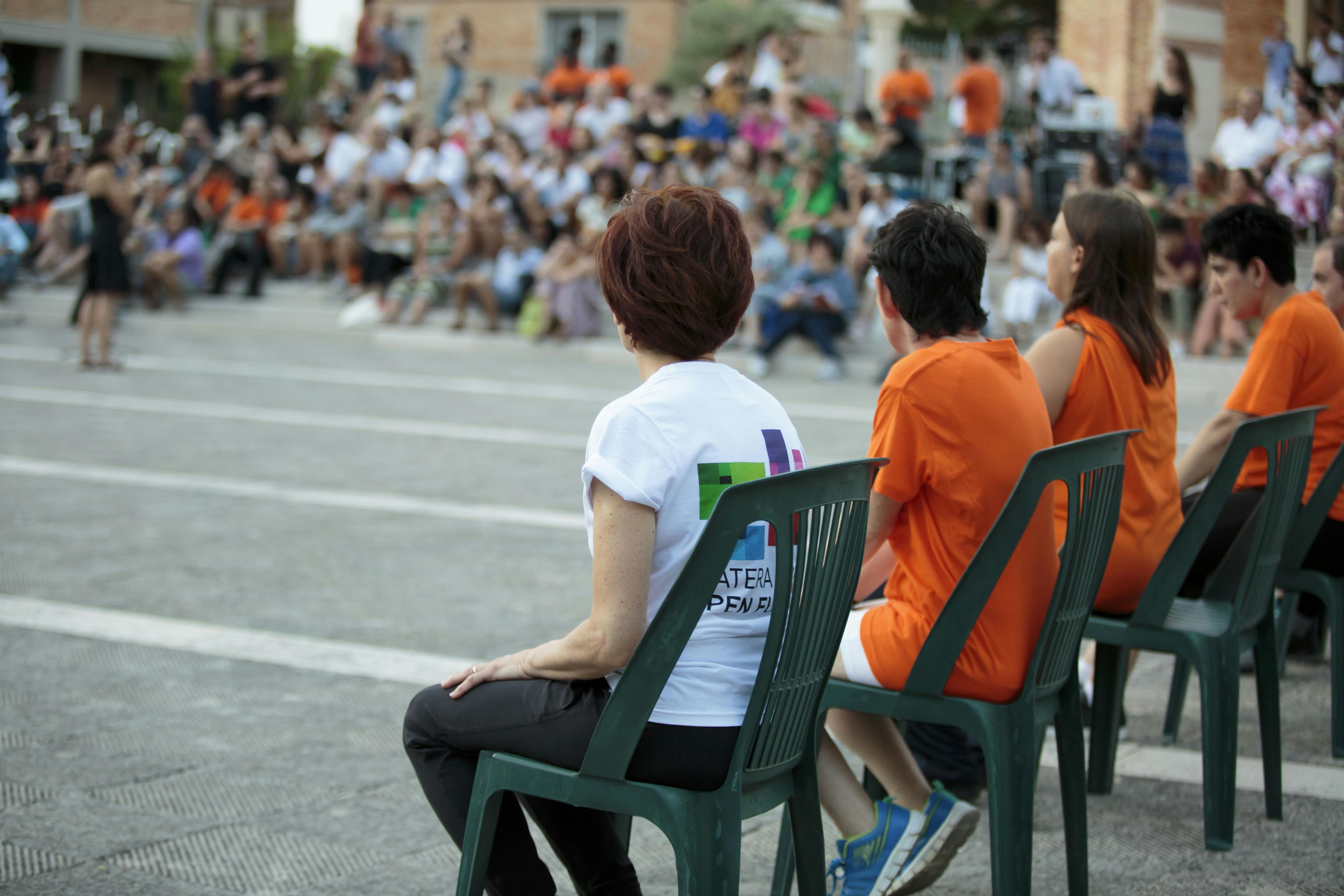 Open air. A group of performers seated before their show. In the background the audience is seated on bleachers.