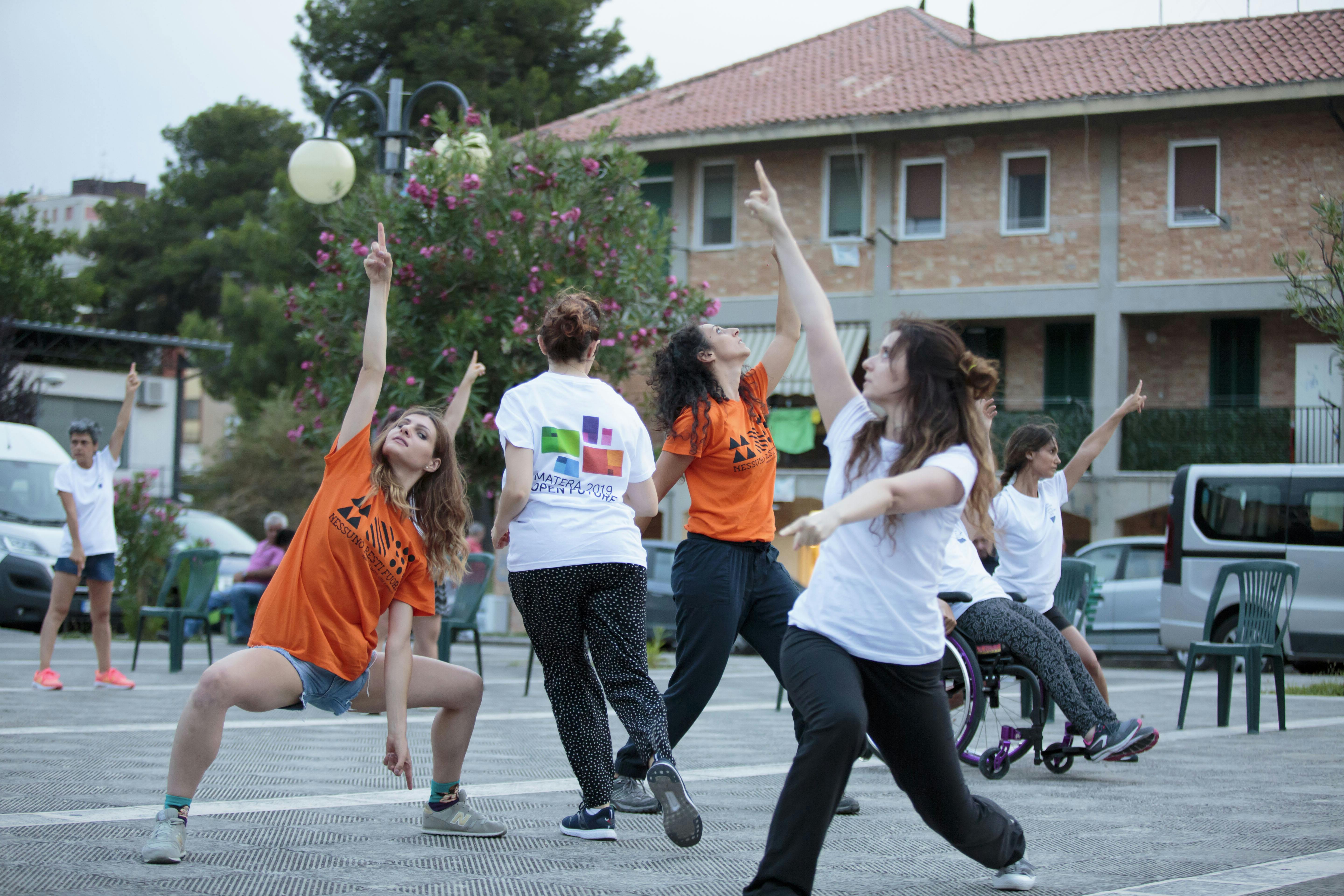 Several dancers with and without disabilities during an outdoor performance. With one arm they point up, with the other toward the ground.