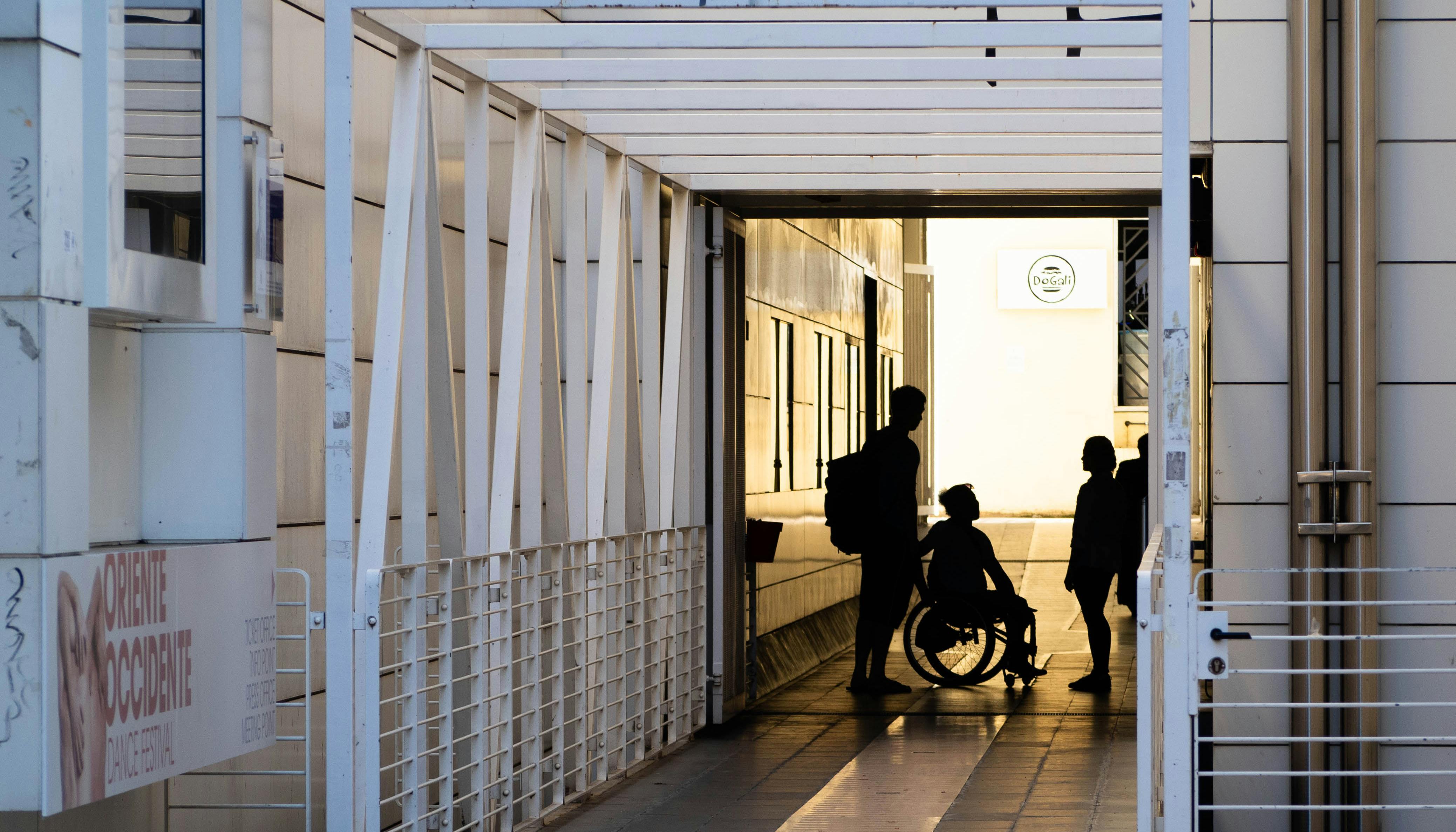 Entrance of Oriente Occidente. The silhouettes of three figures about to enter are shown in backlighting. One of the three is seated in a wheelchair.