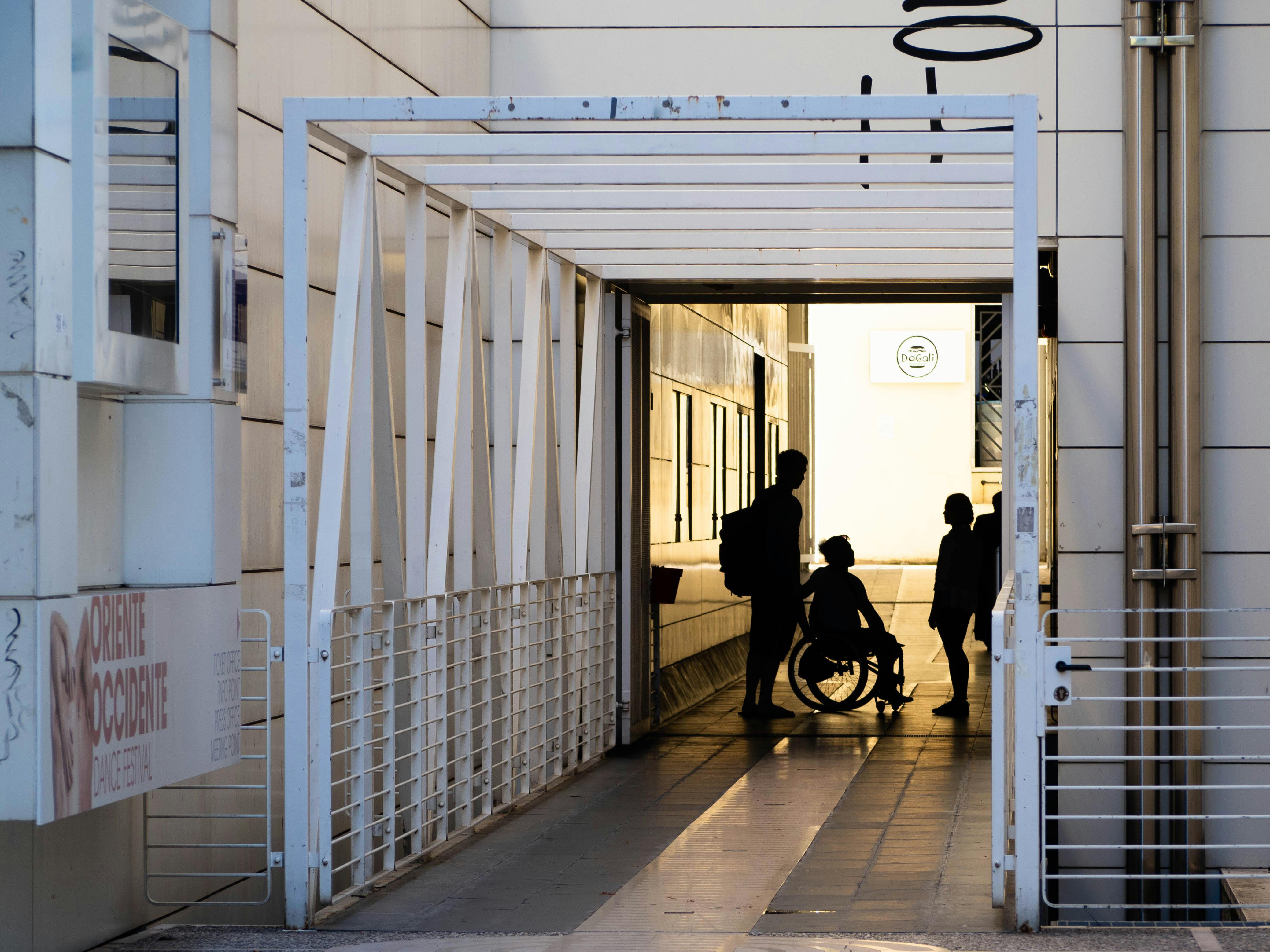 Entrance of Oriente Occidente. The silhouettes of three figures about to enter are shown in backlighting. One of the three is seated in a wheelchair.