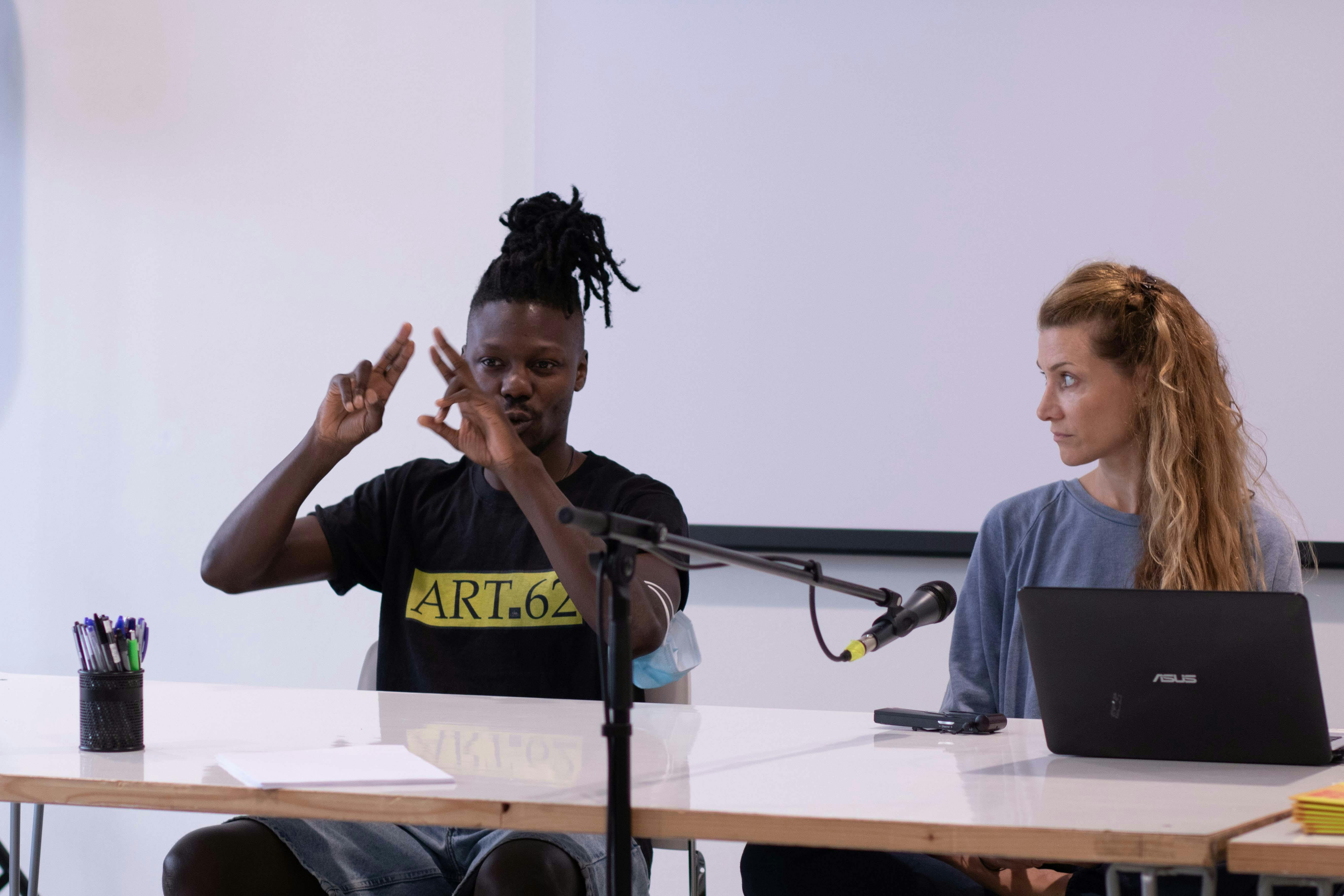 Behind a table, deaf dancer Dodzi Dougban as he signs to the audience in LIS. Next to him, dancer Sarah Bockers is watching.