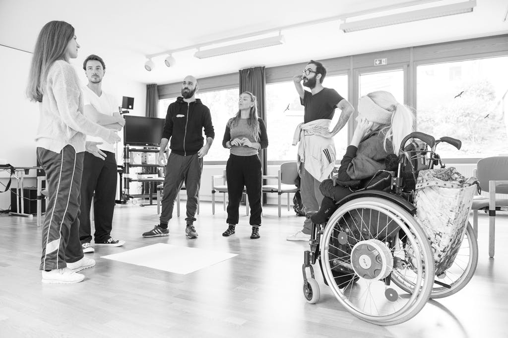 Five able-bodied dancers and a disabled woman in wheelchair. They are standing in a circle and having a discussion.