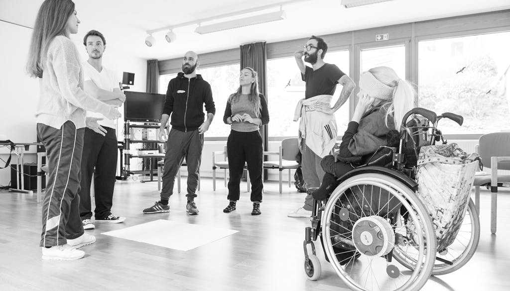 Five able-bodied dancers and a disabled woman in wheelchair. They are standing in a circle and having a discussion.