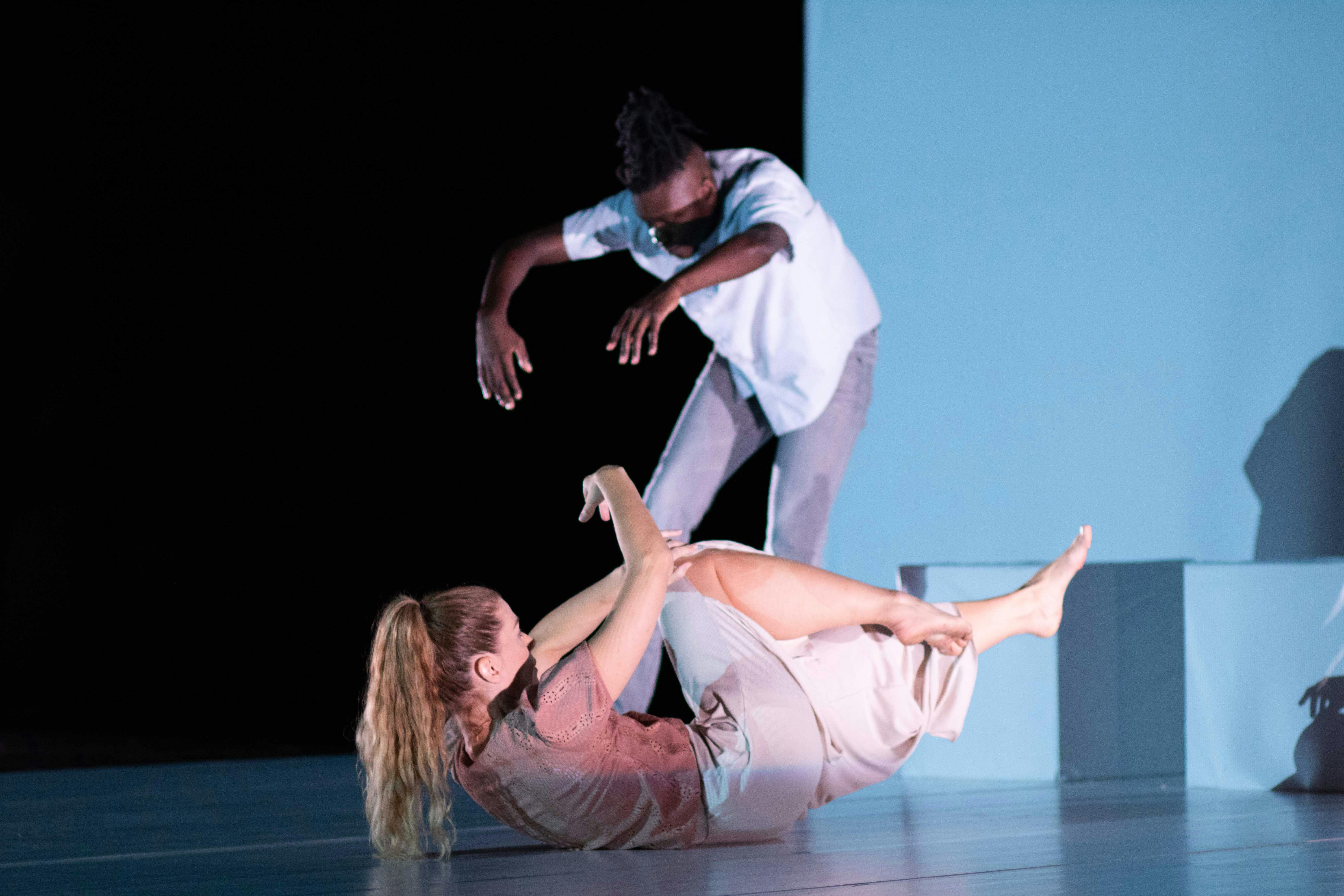 Sarah Bockers and Dodzi Dougban on stage during the preformance Gravity.  She is with her back to the floor, arms and legs raised off the stage; he is standing, with his back in contraction and his arms extended forward.