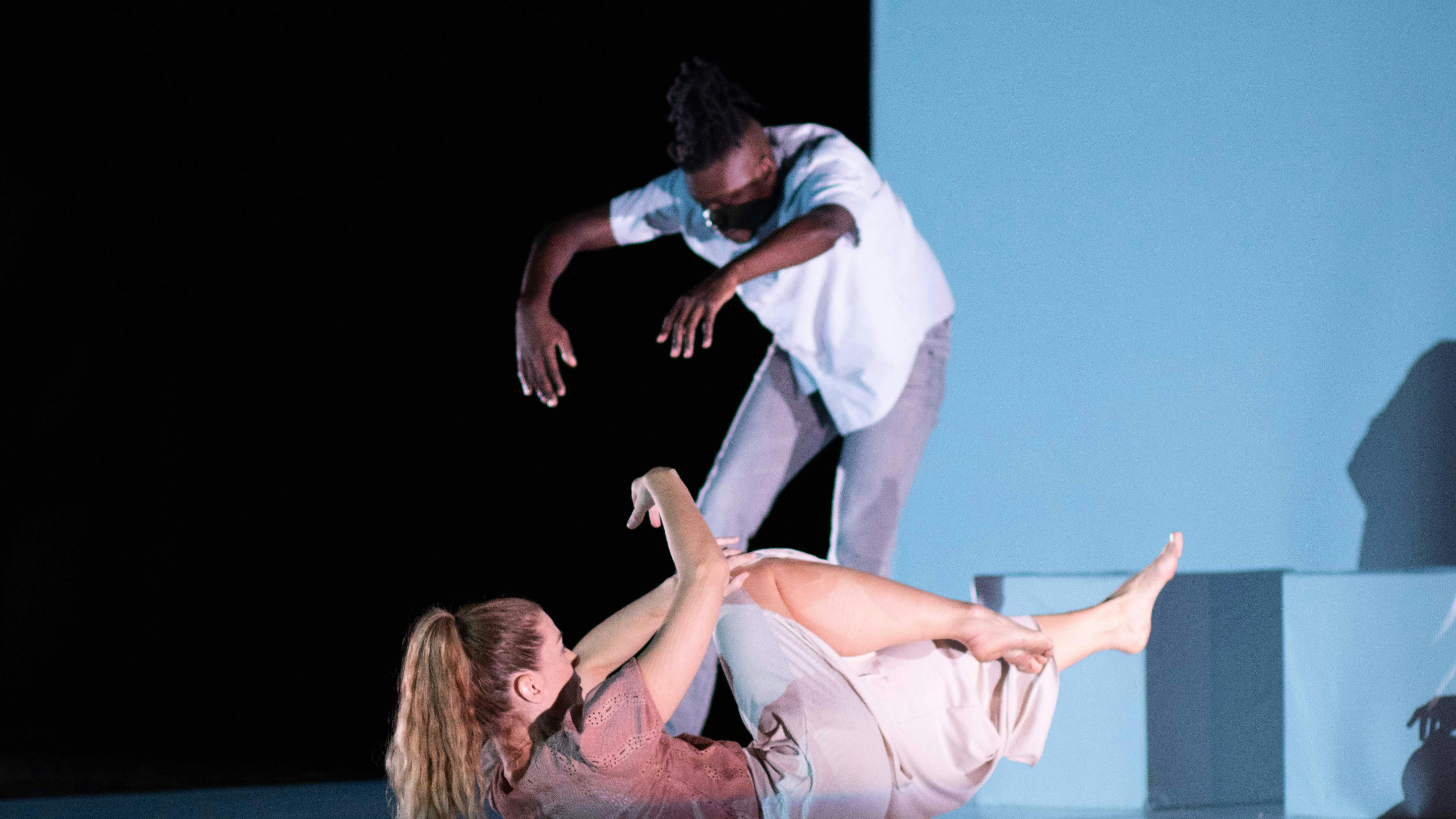 Sarah Bockers and Dodzi Dougban on stage during the preformance Gravity.  She is with her back to the floor, arms and legs raised off the stage; he is standing, with his back in contraction and his arms extended forward.