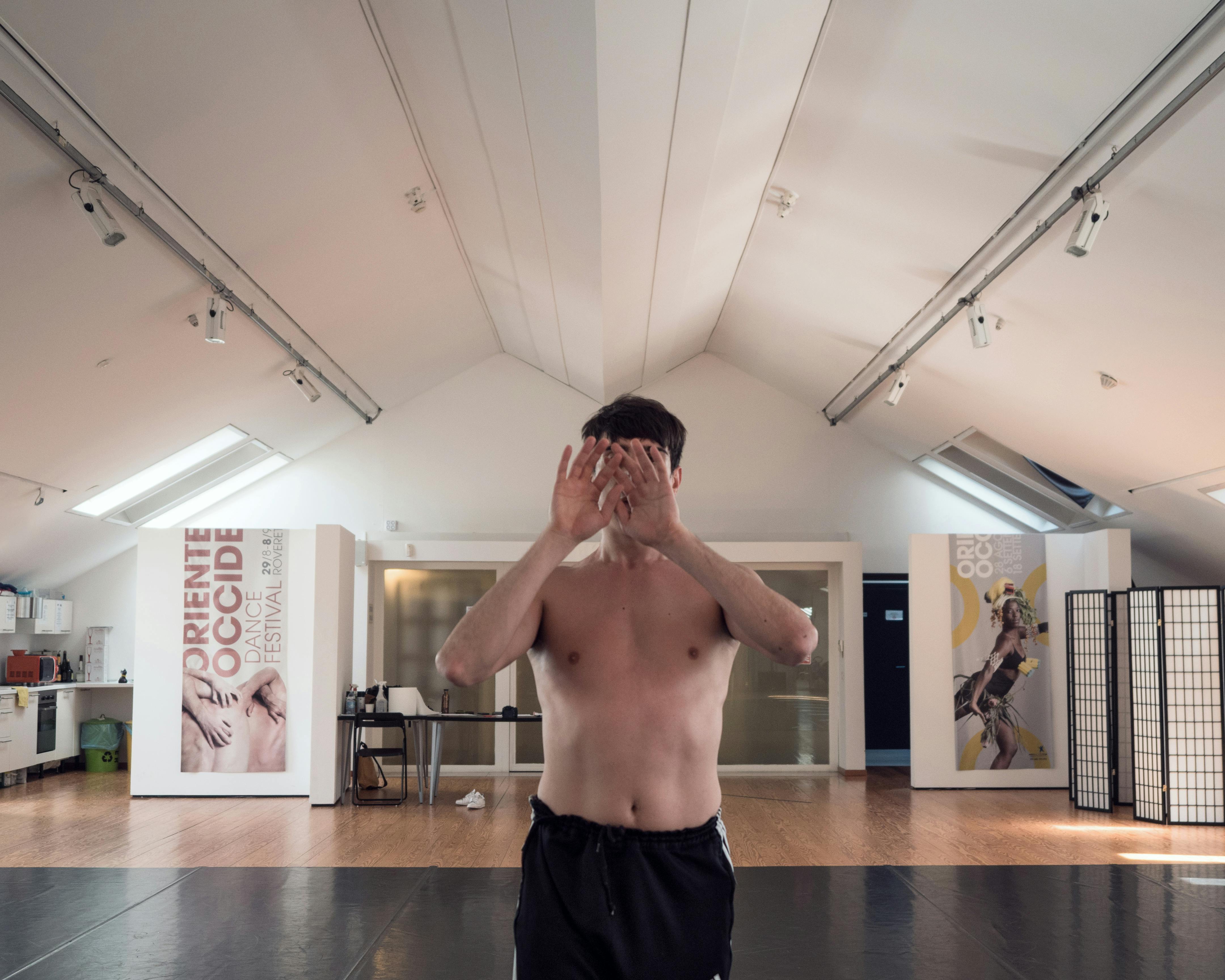 Il danzatore Cristian Cucco a torso nudo nel centro dello Studio si copre il volto con le mani rivolgendo i palmi verso l'obiettivo