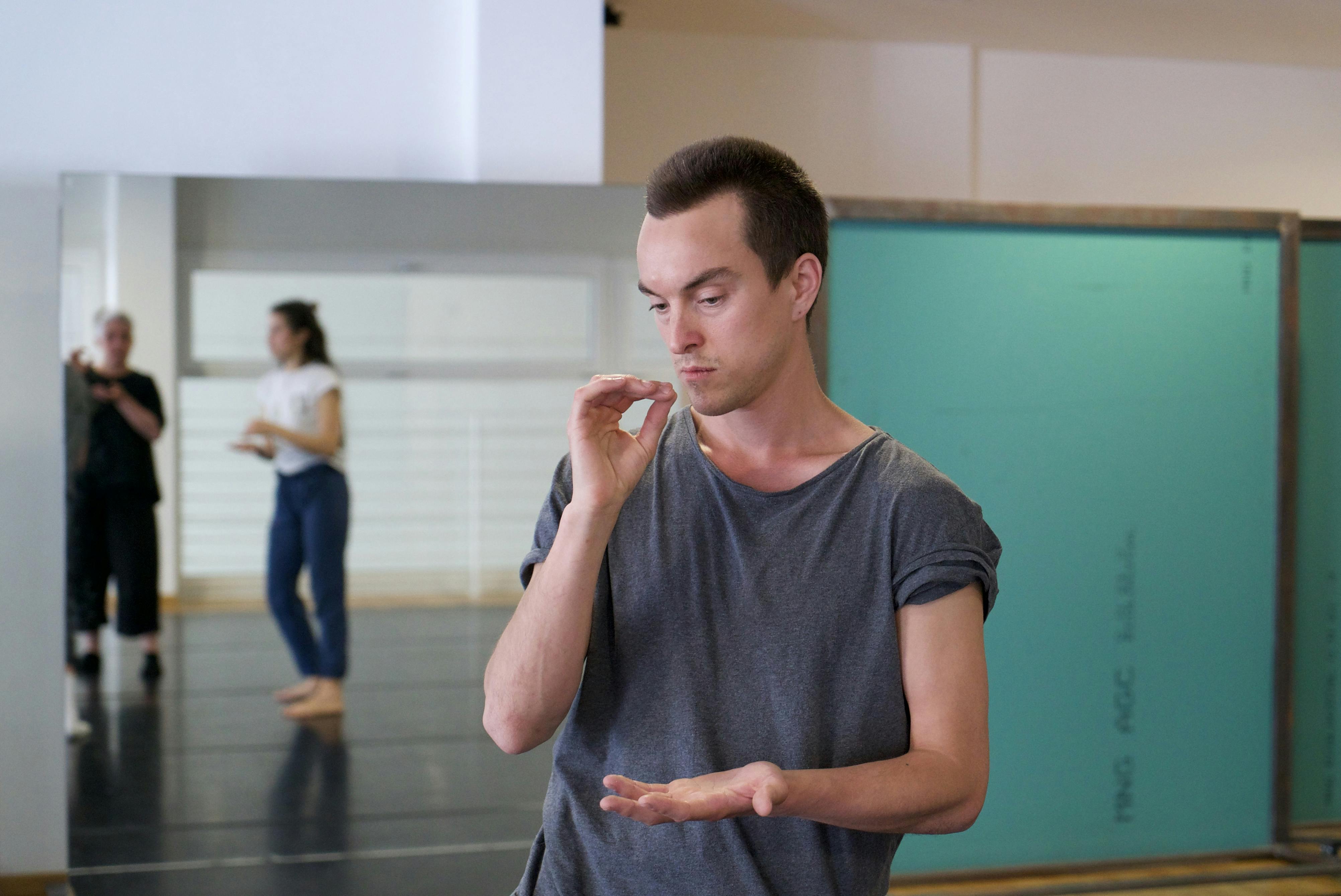 Sindri Runudde rehearsing in the Studio with a few local volunteers. The artist is shown frontally; behind him, reflected in the mirror, two women imitate his gesture.