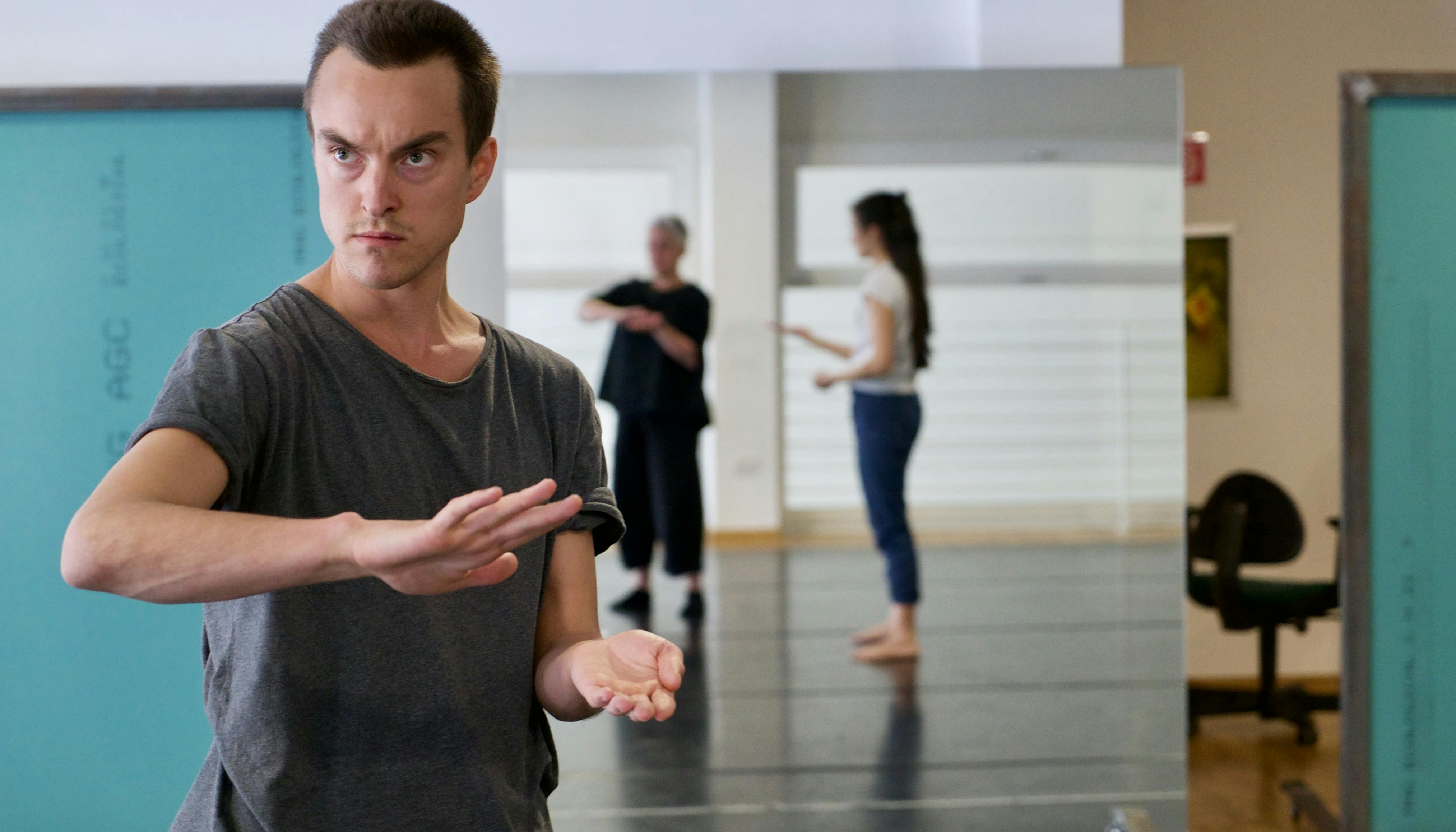 Sindri Runudde rehearsing in the Studio together with a few local volunteers. The artist is shown frontally; behind him, reflected in the mirror, two women imitate his gesture.