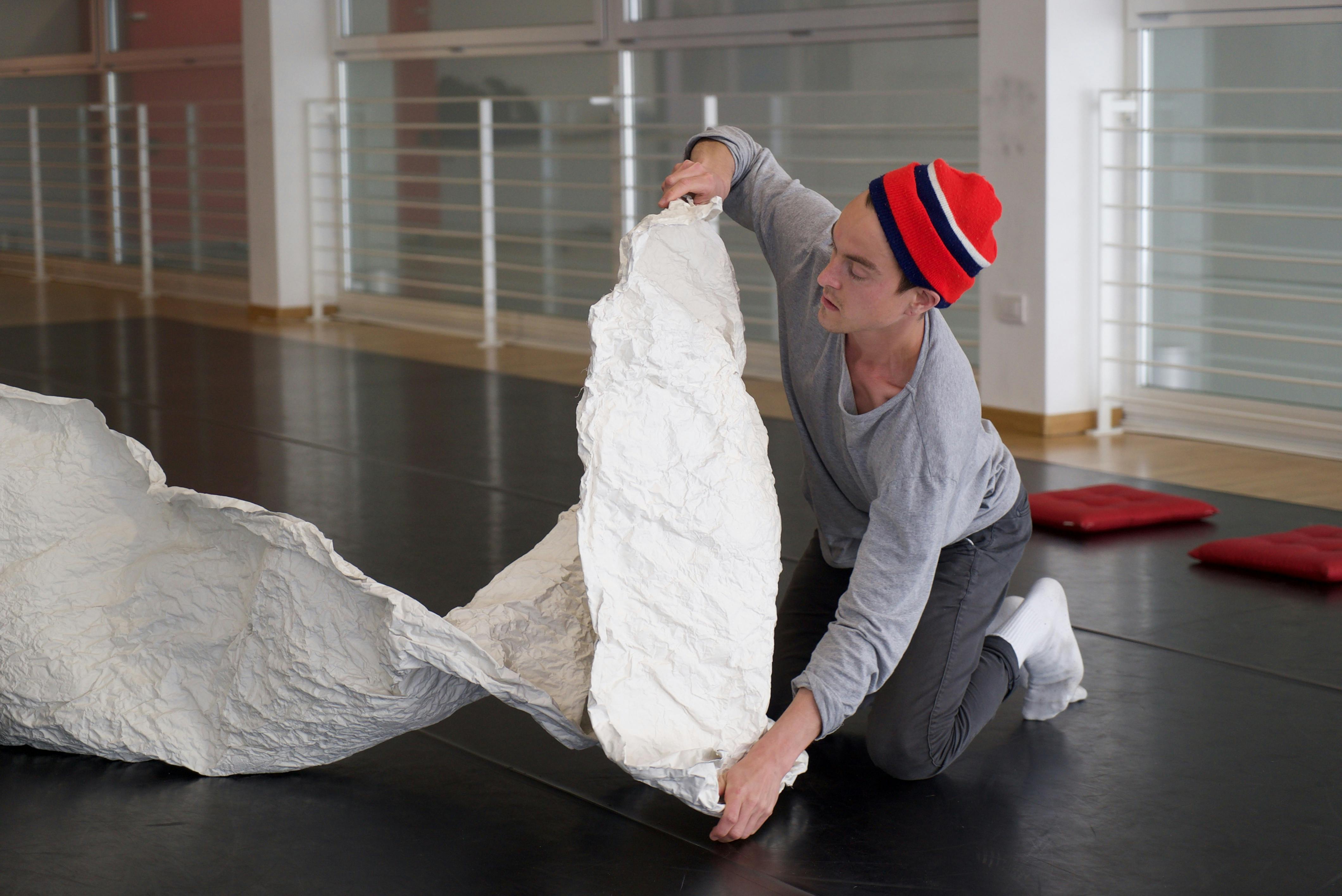 Sindri Runudde during an open rehearsal in the Studio. He is wearing a colorful woolen cap, is kneeled on the ground; he holds the edge of a long strip of crumpled white paper in his hands.