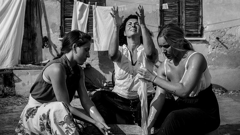 Gocce by Federica Loredan Three women dressed in summer clothes are washing clothes in a basin. They are arranged in a semi-circle, the central one with her eyes and hands turned towards the sky.