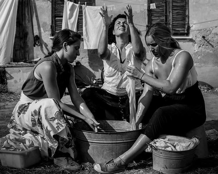 Three women dressed in summer clothes are washing clothes in a basin. They are arranged in a semi-circle, the central one with her eyes and hands turned towards the sky.
