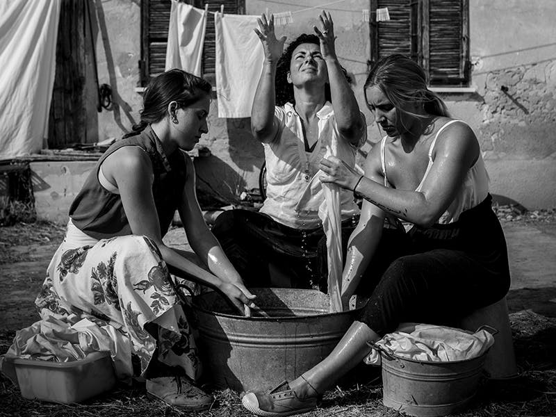 Three women dressed in summer clothes are washing clothes in a basin. They are arranged in a semi-circle, the central one with her eyes and hands turned towards the sky.