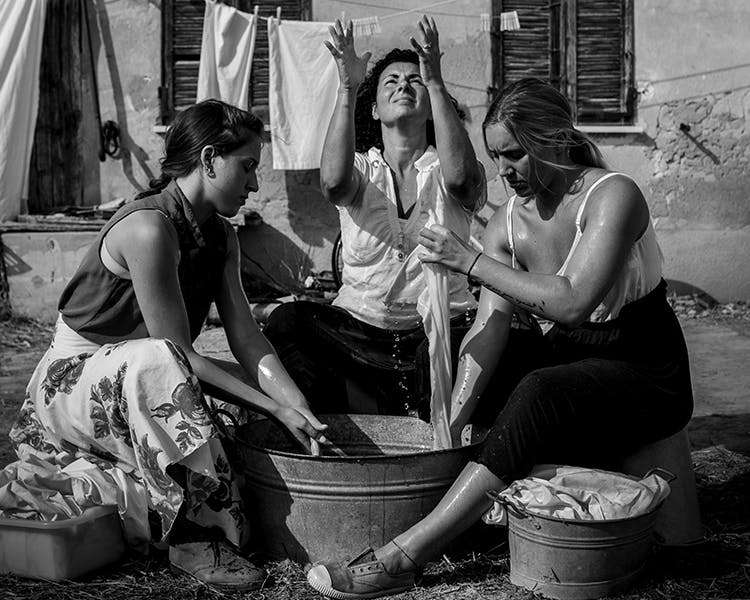 Tre donne vestite con abiti estivi lavano i panni in un catino. Sono sistemate a semicercio, quella centrale ha lo sguardo e le mani rivolti verso il cielo