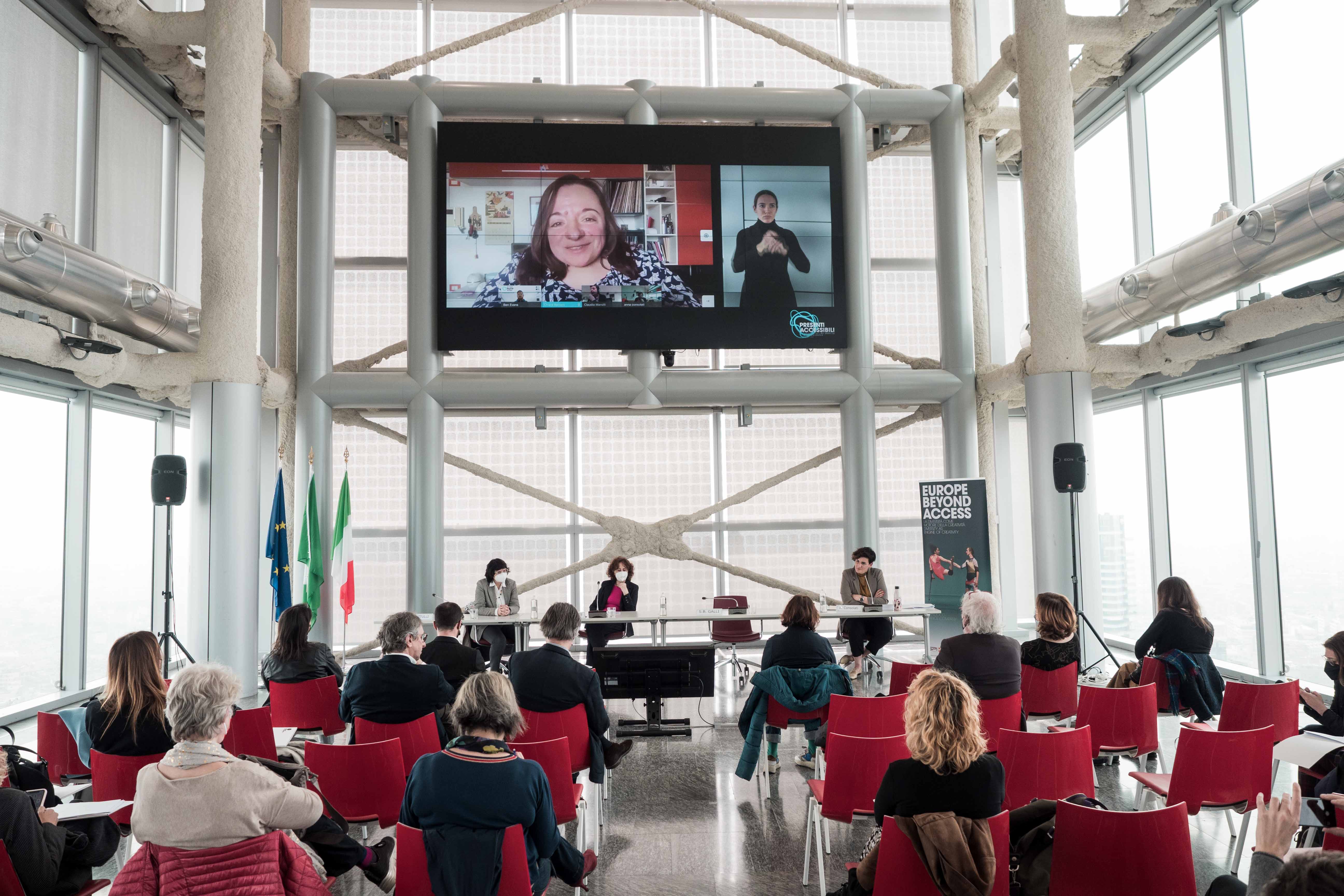 The audience, seen from behind, looks towards the speakers' table at the Presenti accessibili press conference. Above, a large screen shows a remote speaker and LIS translator.