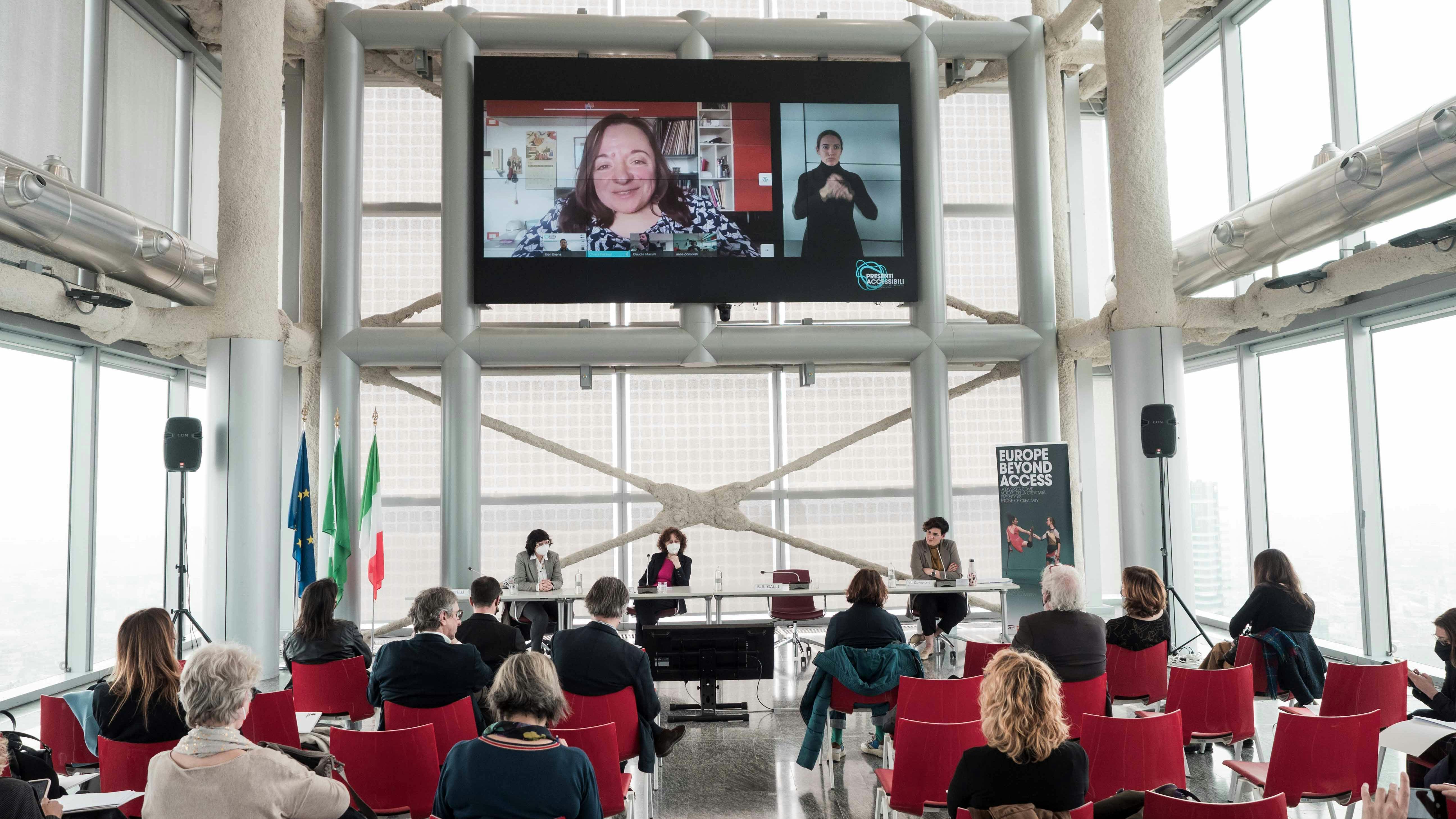 Presenti accessibili, press conference The audience, seen from behind, looks towards the speakers' table at the Presenti accessibili press conference. Above, a large screen shows a remote speaker and LIS translator.