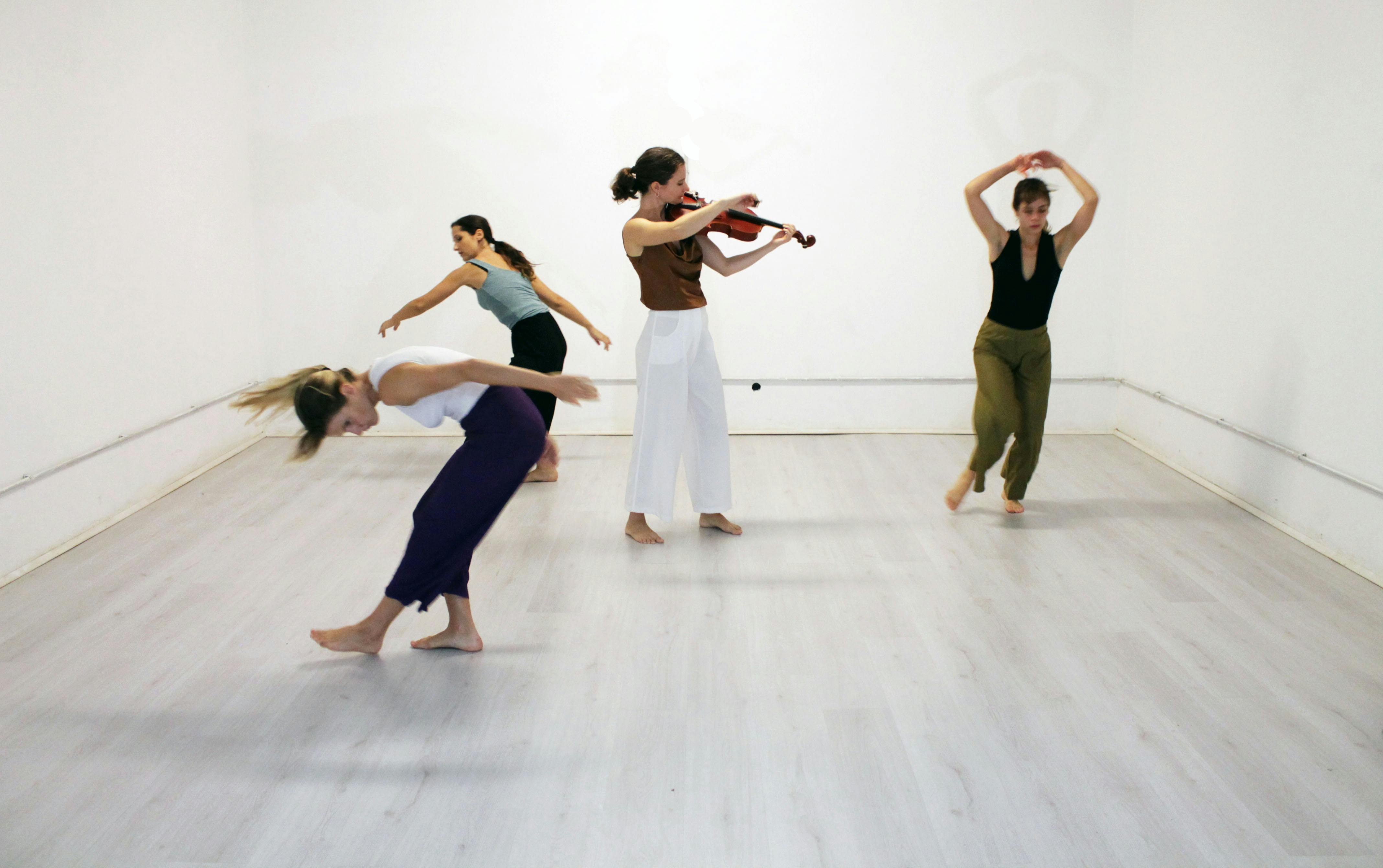 A violinist in the center of the room plays and three performers dance around her
