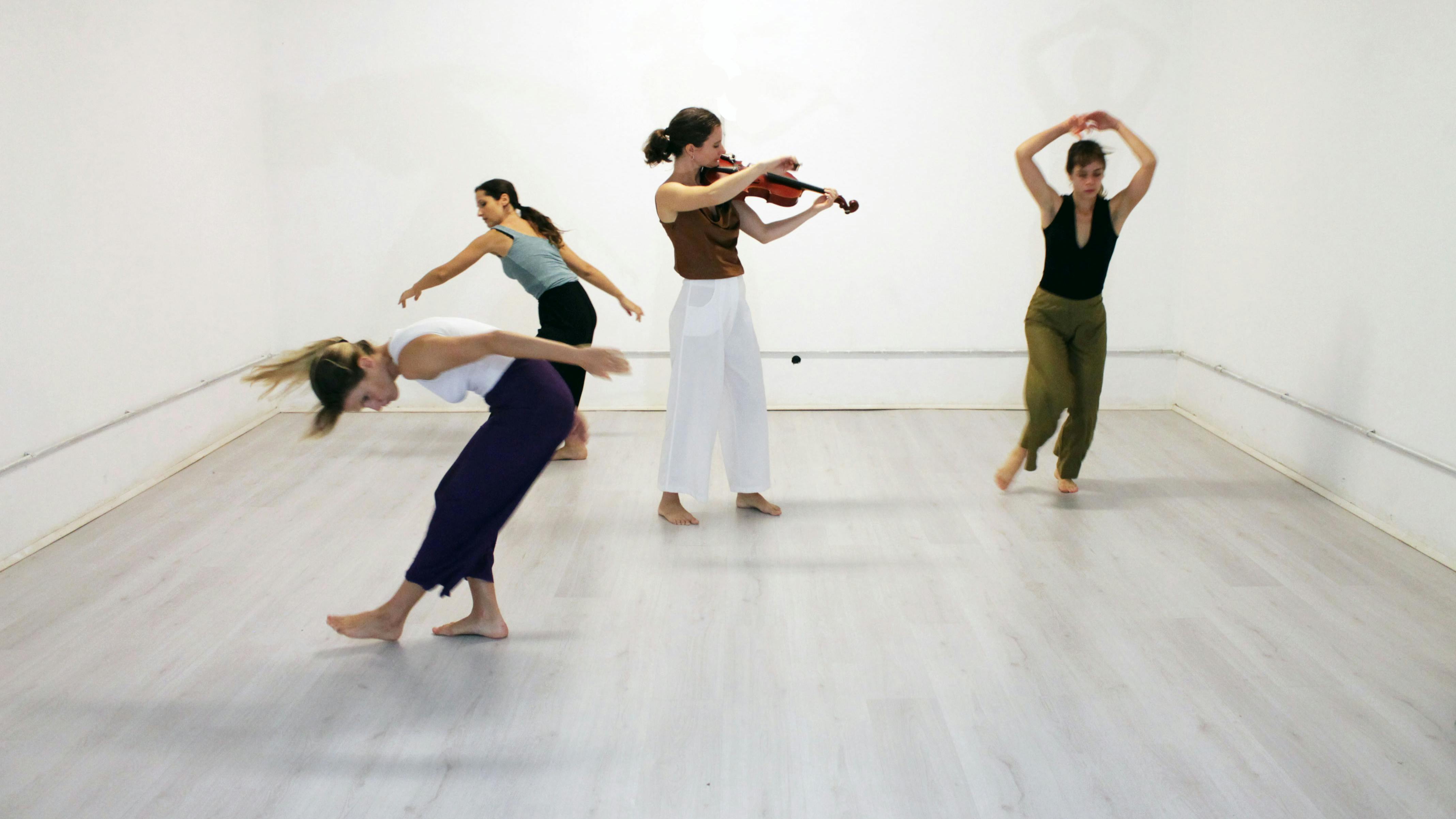 A violinist in the center of the room plays and three performers dance around her
