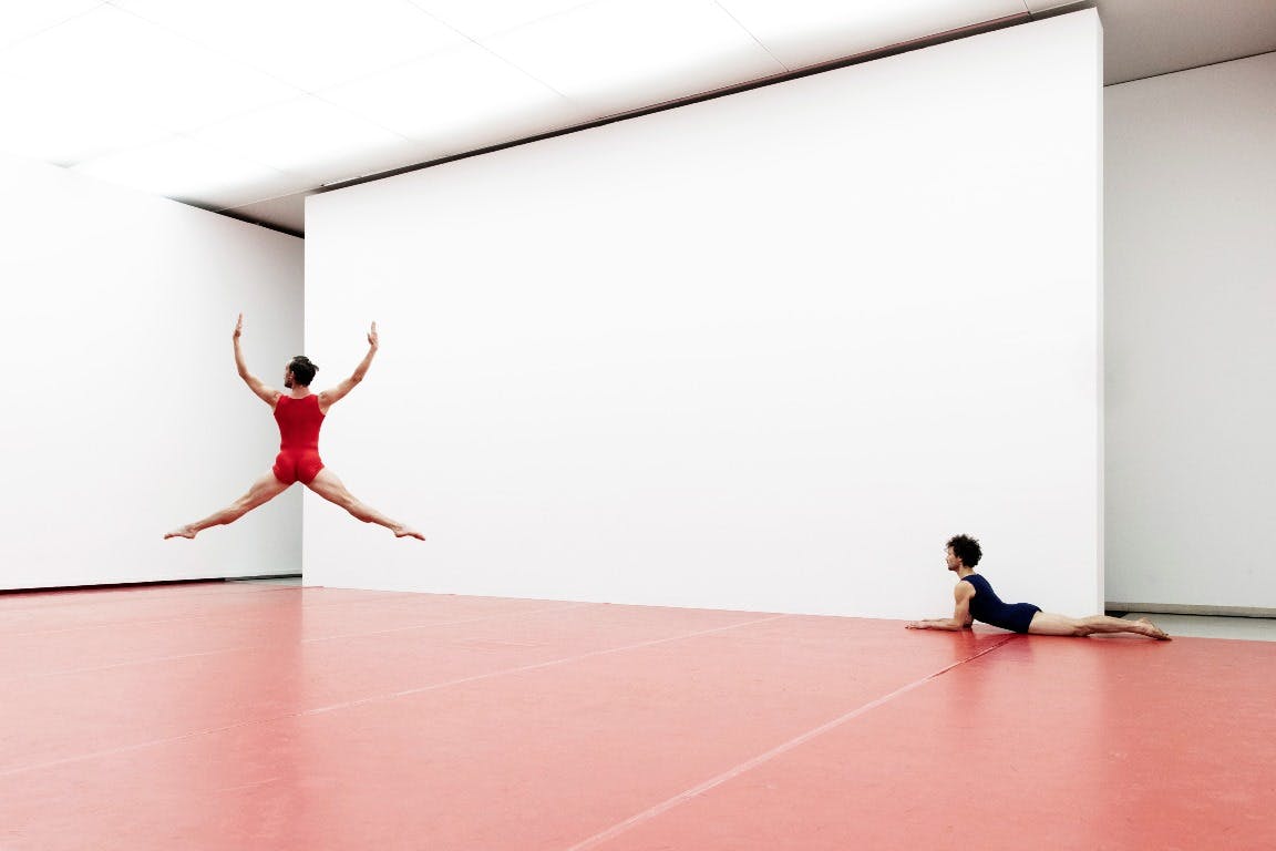 One dancer in red jumpsuit performs a jump while the other is lying down
