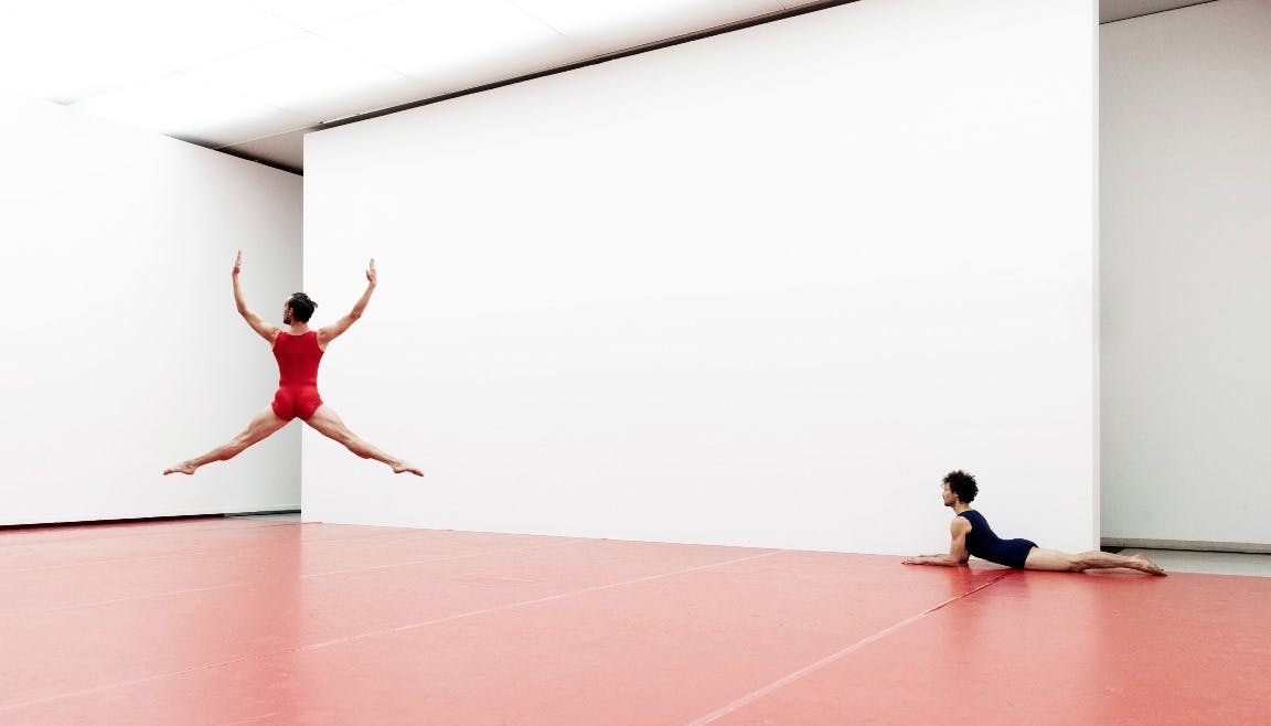 One dancer in red jumpsuit performs a jump while the other is lying down