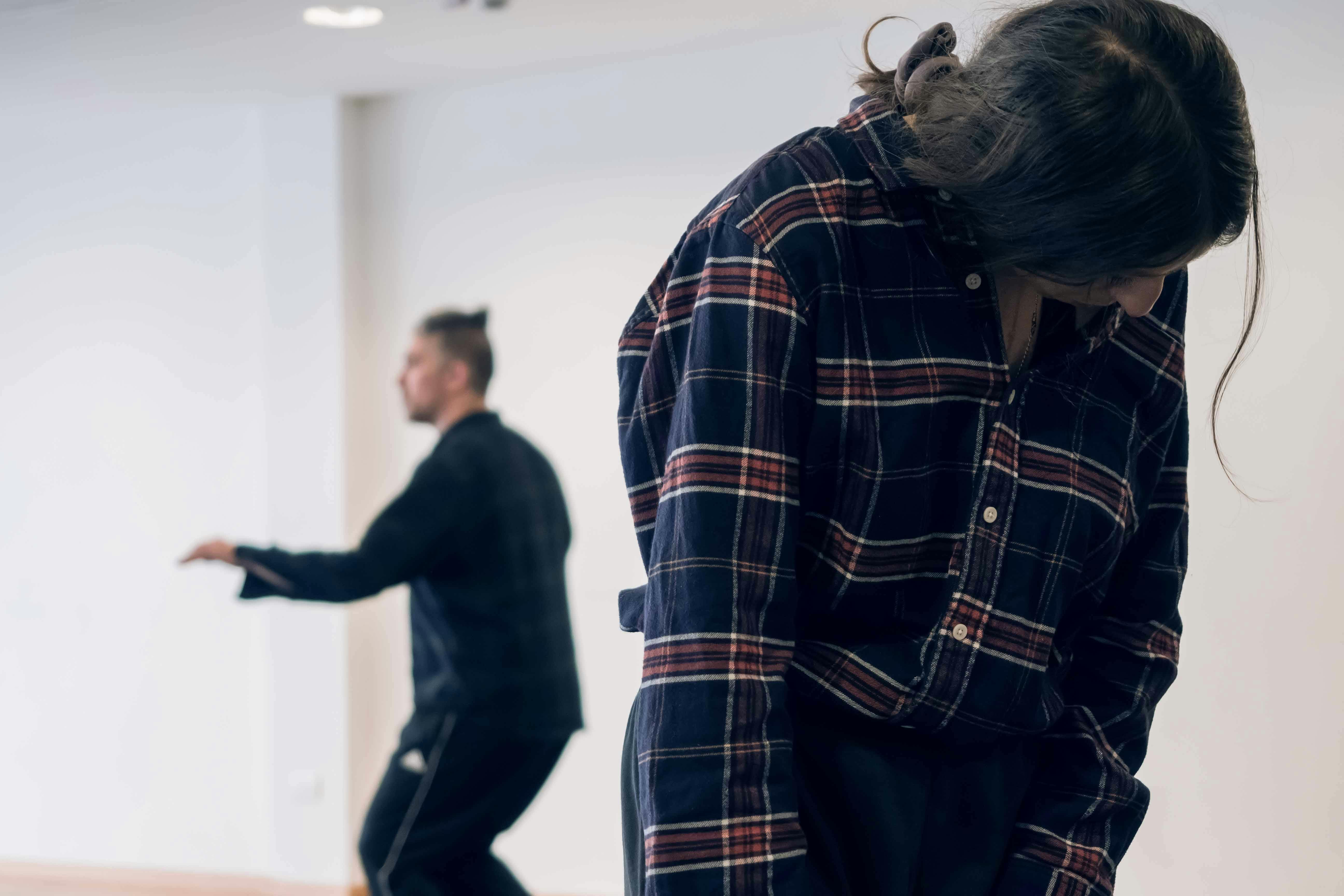 In primo piano una delle interpreti del progetto che guarda verso in basso, sullo sfondo il coreografo con un braccio allungato in avanti 