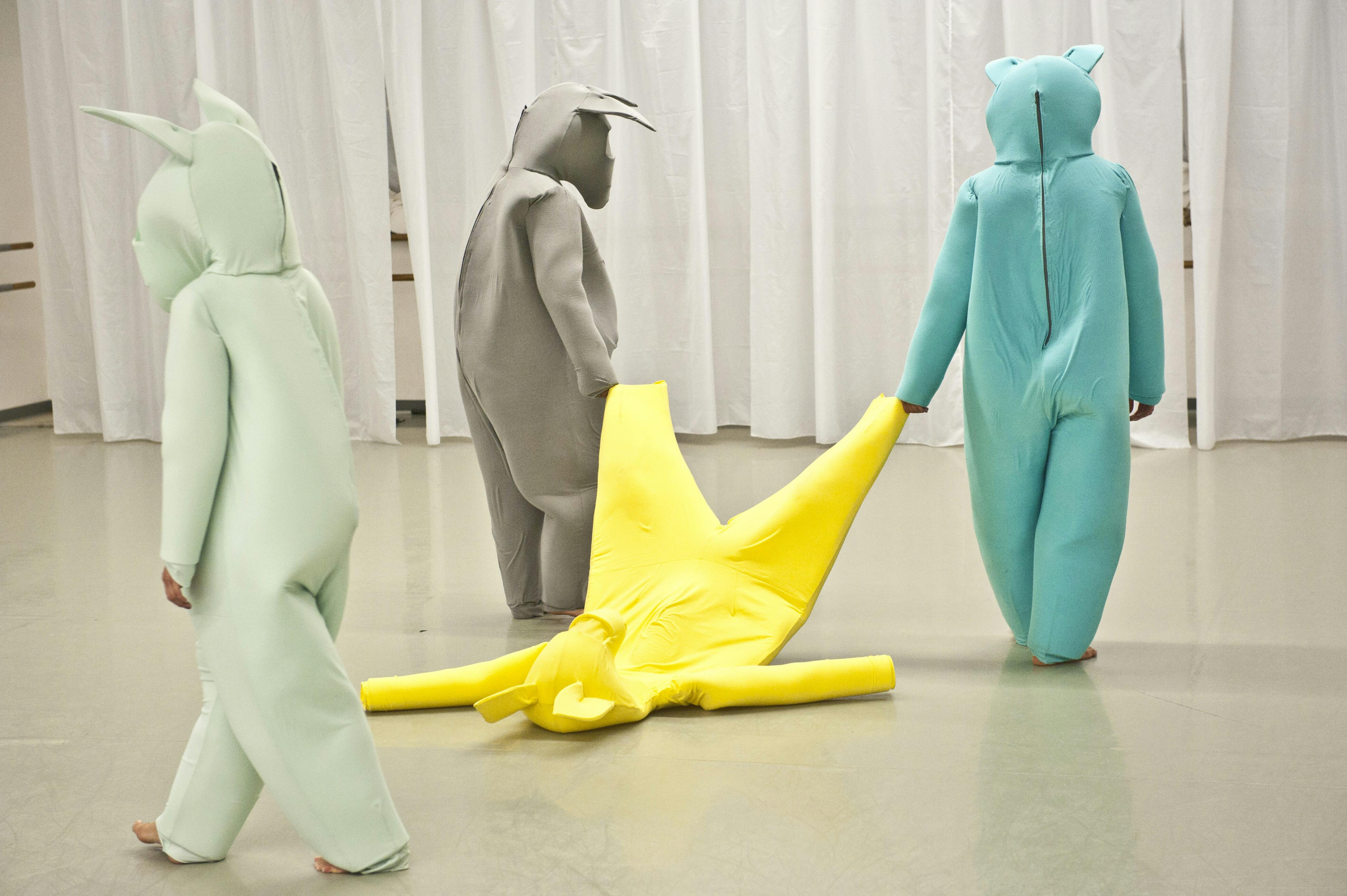 Three dancers in colorful jumpsuits drag a fourth suit