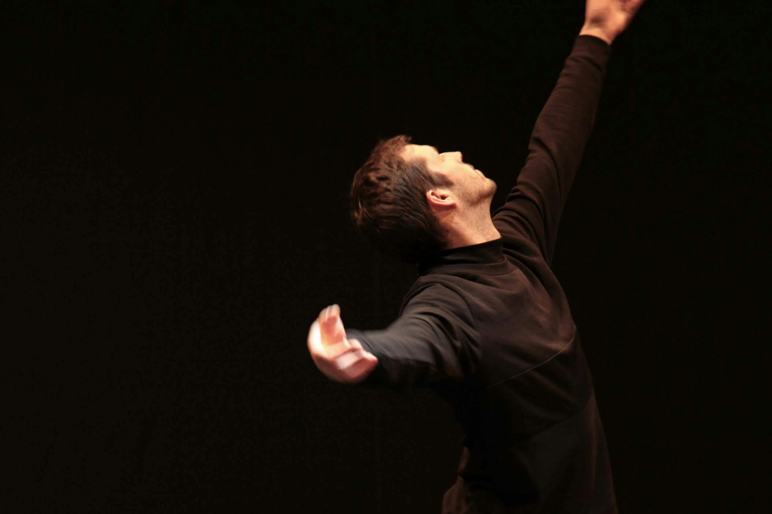 il danzatore vestito di nero, su sfondo nero ha il braccio sinistro alzato e lo sguardo alla mano