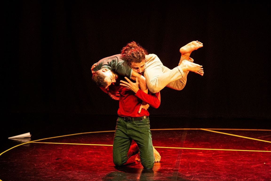 Un performer inginocchiato porta sulle spalle un secondo interprete