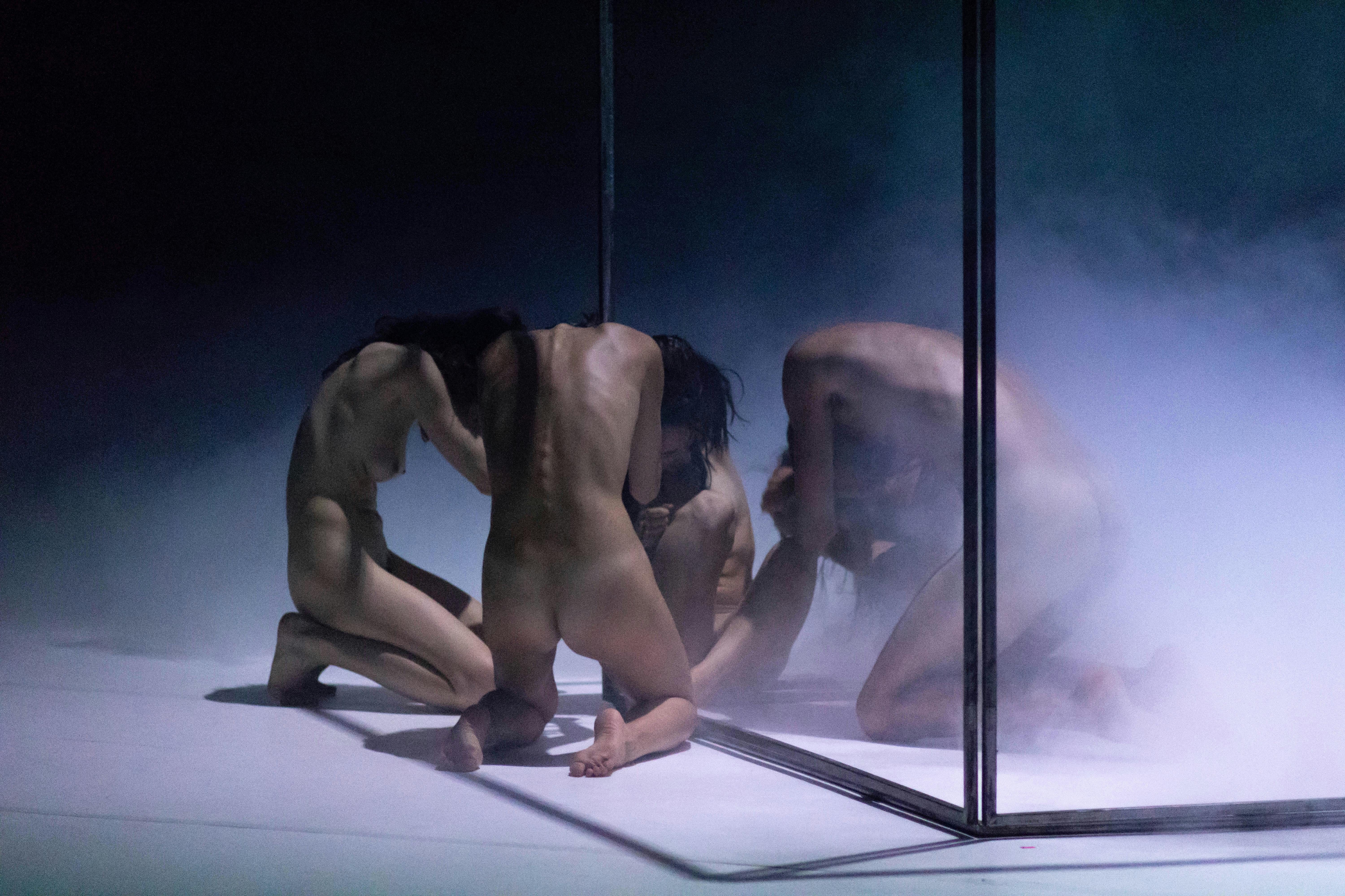 Five dancers kneeling on the stage, naked. They hold their heads in their hands. Smoke mists the scene.