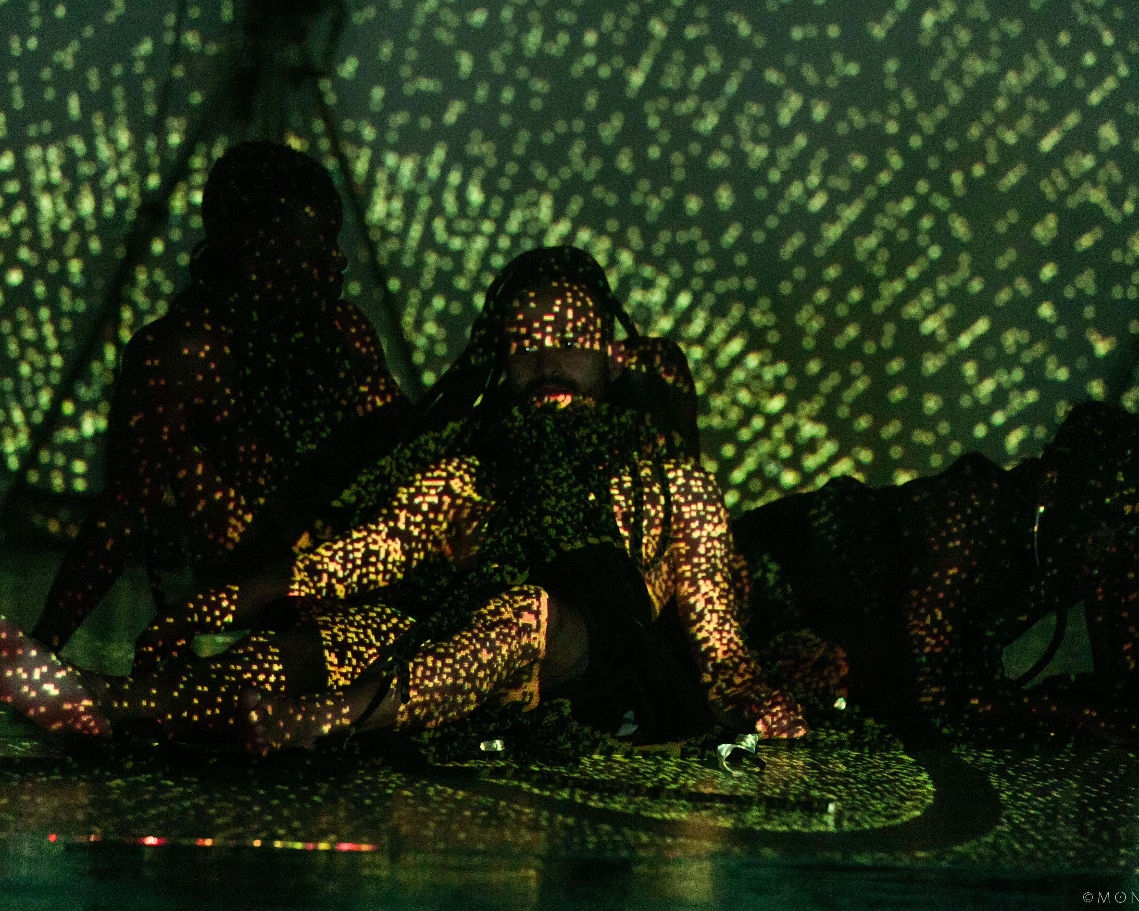 Tre danzatori seduti illuminati da luci verdi e gialle