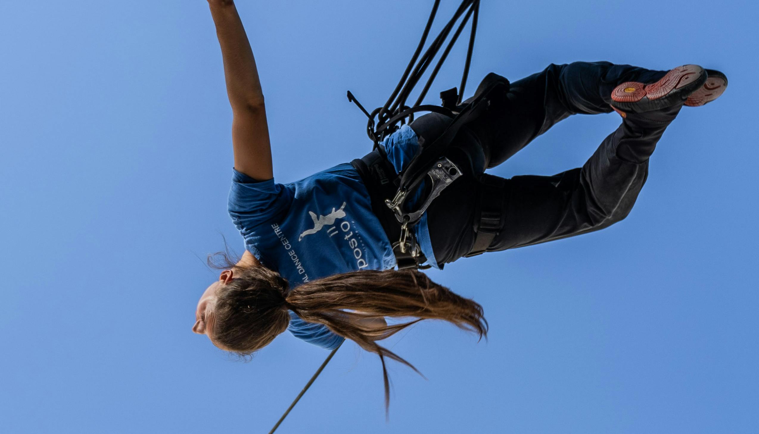 A dancer wearing a harness and hanging by a thread stands out against the sky with her back arched and her right arm open. Her long hair are laced in a tail