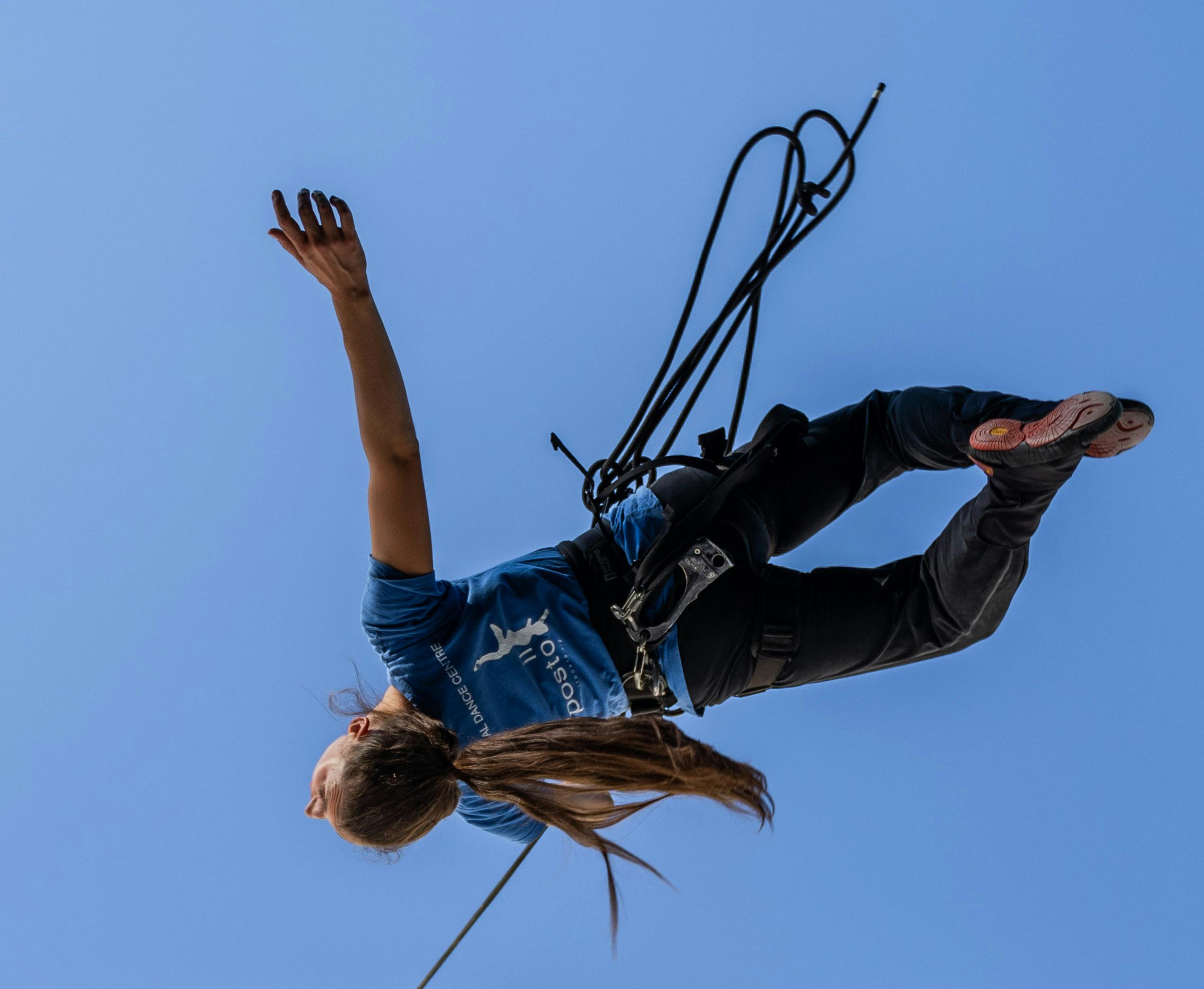 A dancer wearing a harness and hanging by a thread stands out against the sky with her back arched and her right arm open. Her long hair are laced in a tail