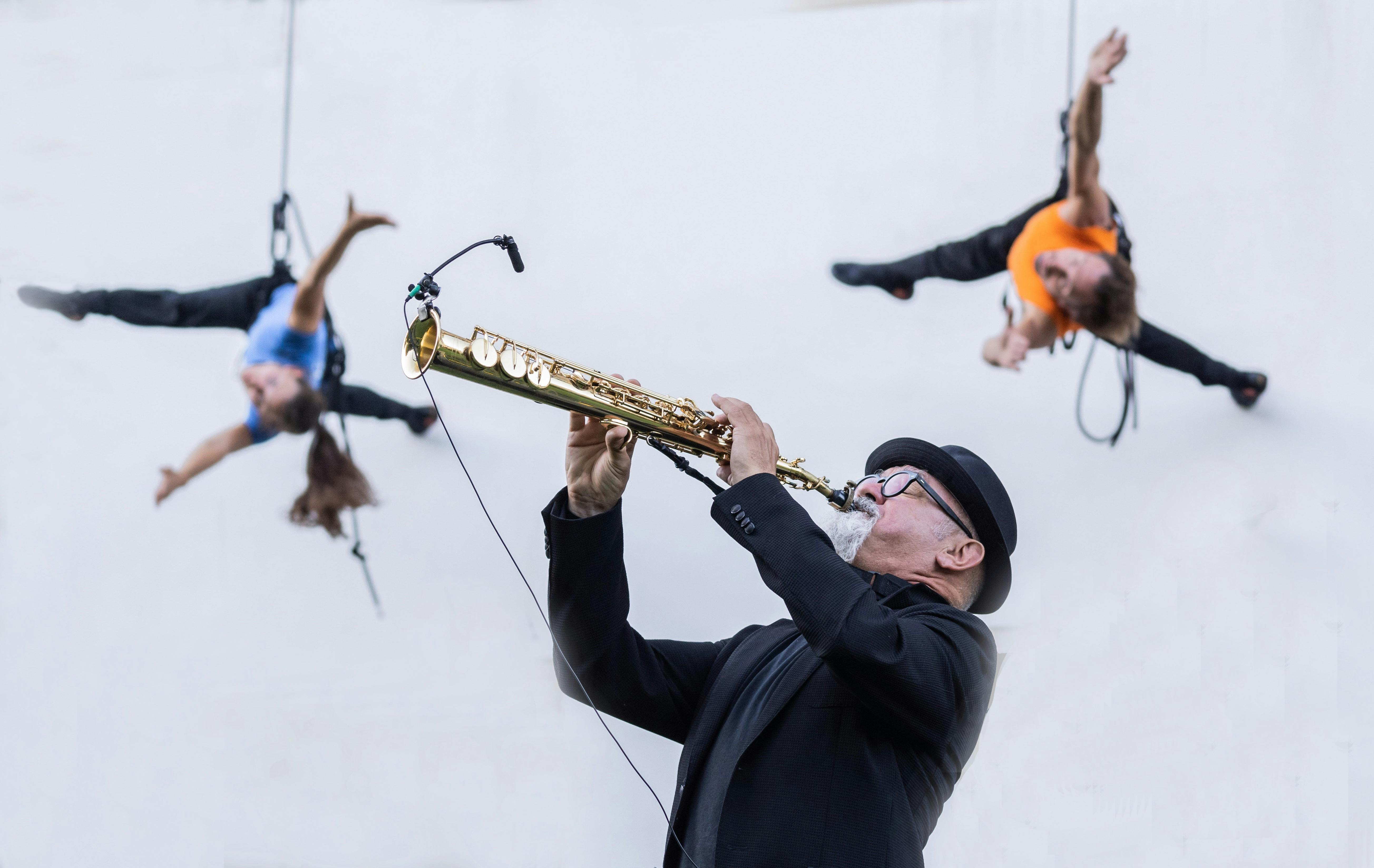 In the foreground a musician plays a trumpet. He is wearing a jacket, a bowler hat and black spectacles. In the background, two performers dance harnessed on a grey wall, a man in an orange T-shirt and a woman with long hair pulled back in a ponytail and a blue T-shirt.