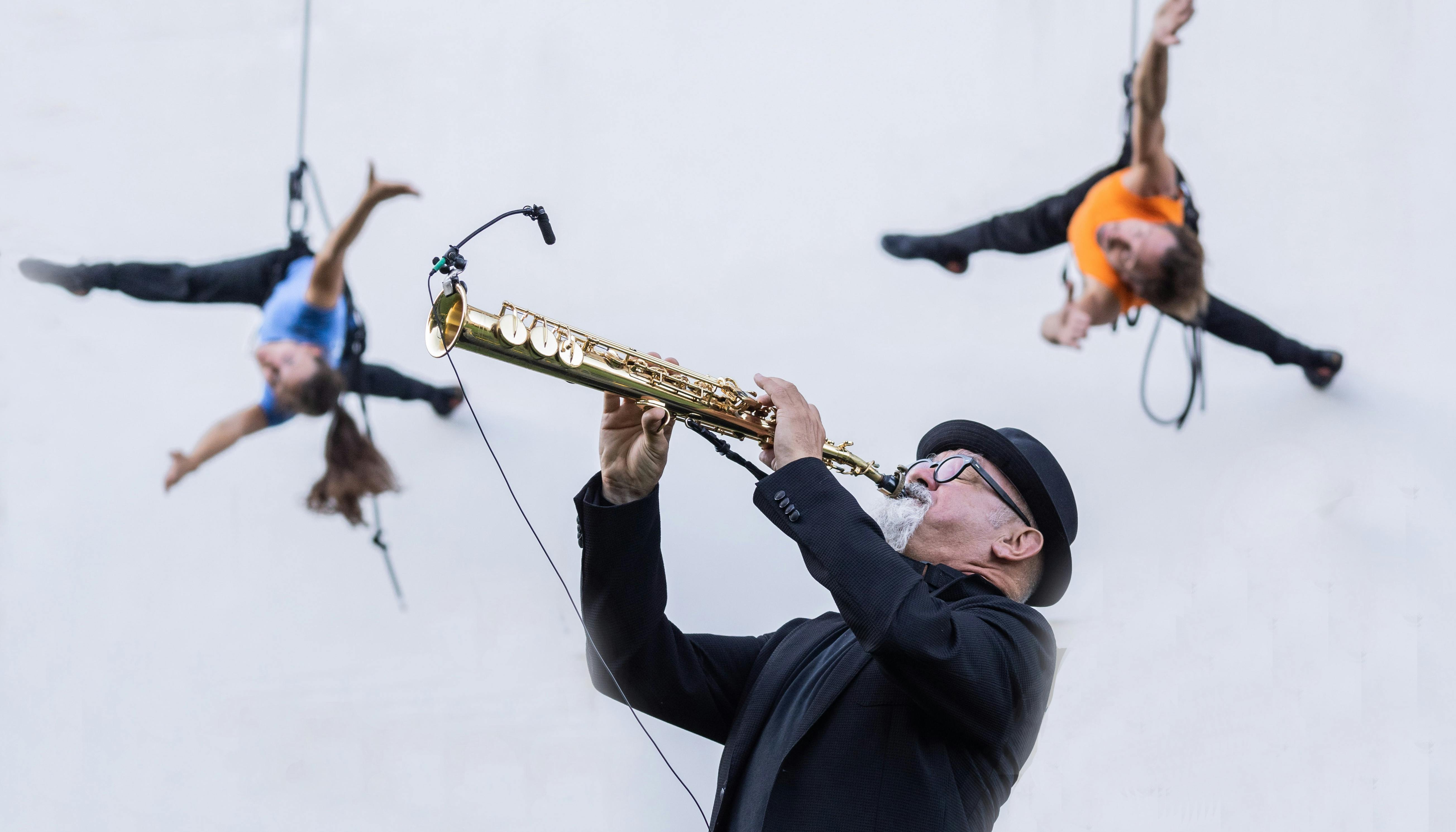 In the foreground a musician plays a trumpet. He is wearing a jacket, a bowler hat and black spectacles. In the background, two performers dance harnessed on a grey wall, a man in an orange T-shirt and a woman with long hair pulled back in a ponytail and a blue T-shirt.