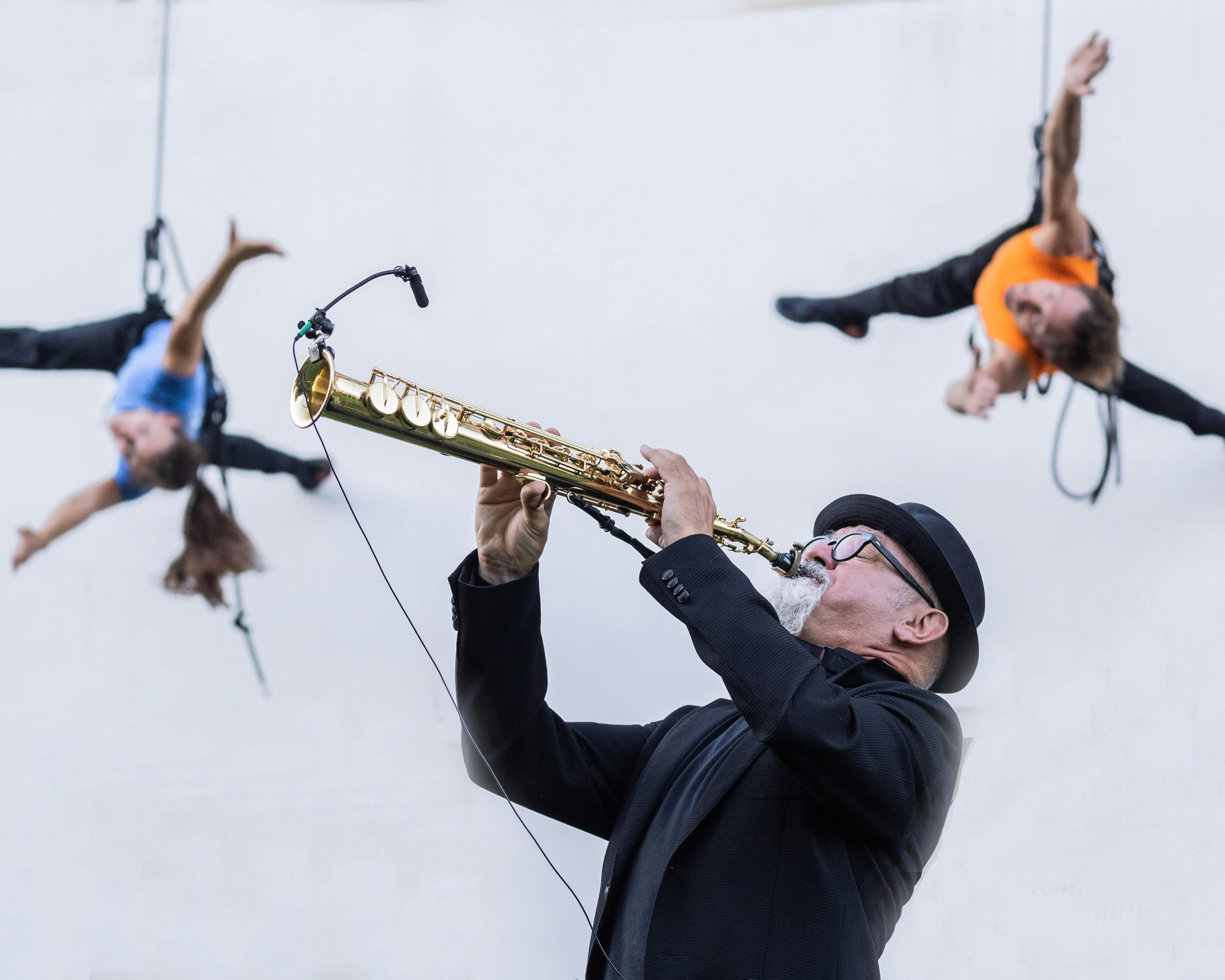 In the foreground a musician plays a trumpet. He is wearing a jacket, a bowler hat and black spectacles. In the background, two performers dance harnessed on a grey wall, a man in an orange T-shirt and a woman with long hair pulled back in a ponytail and a blue T-shirt.