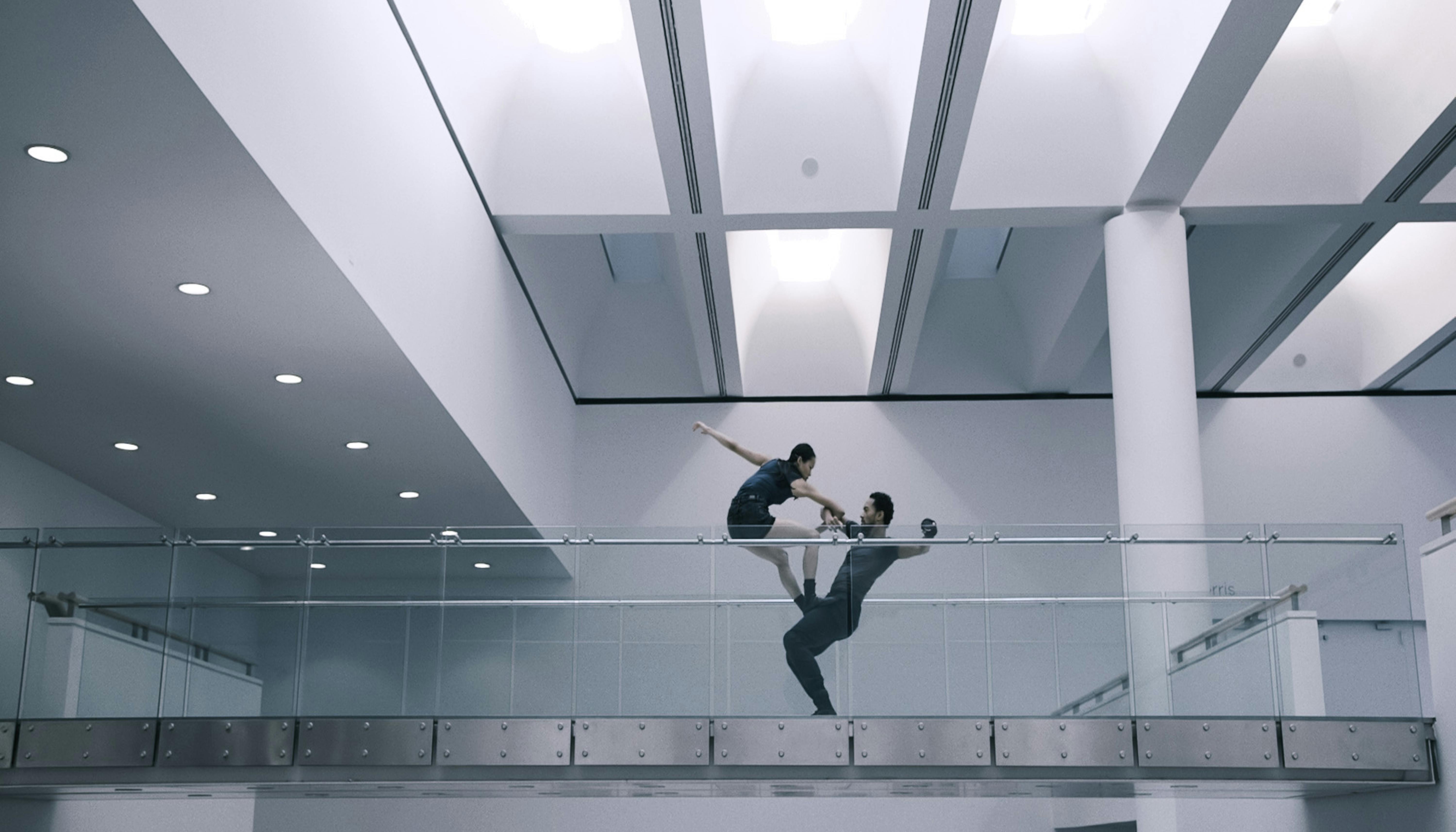 Un danzatore sostiene sulle gambe una danzatrice all'interno di uno spazio museale. I colori sono bianco e grigio