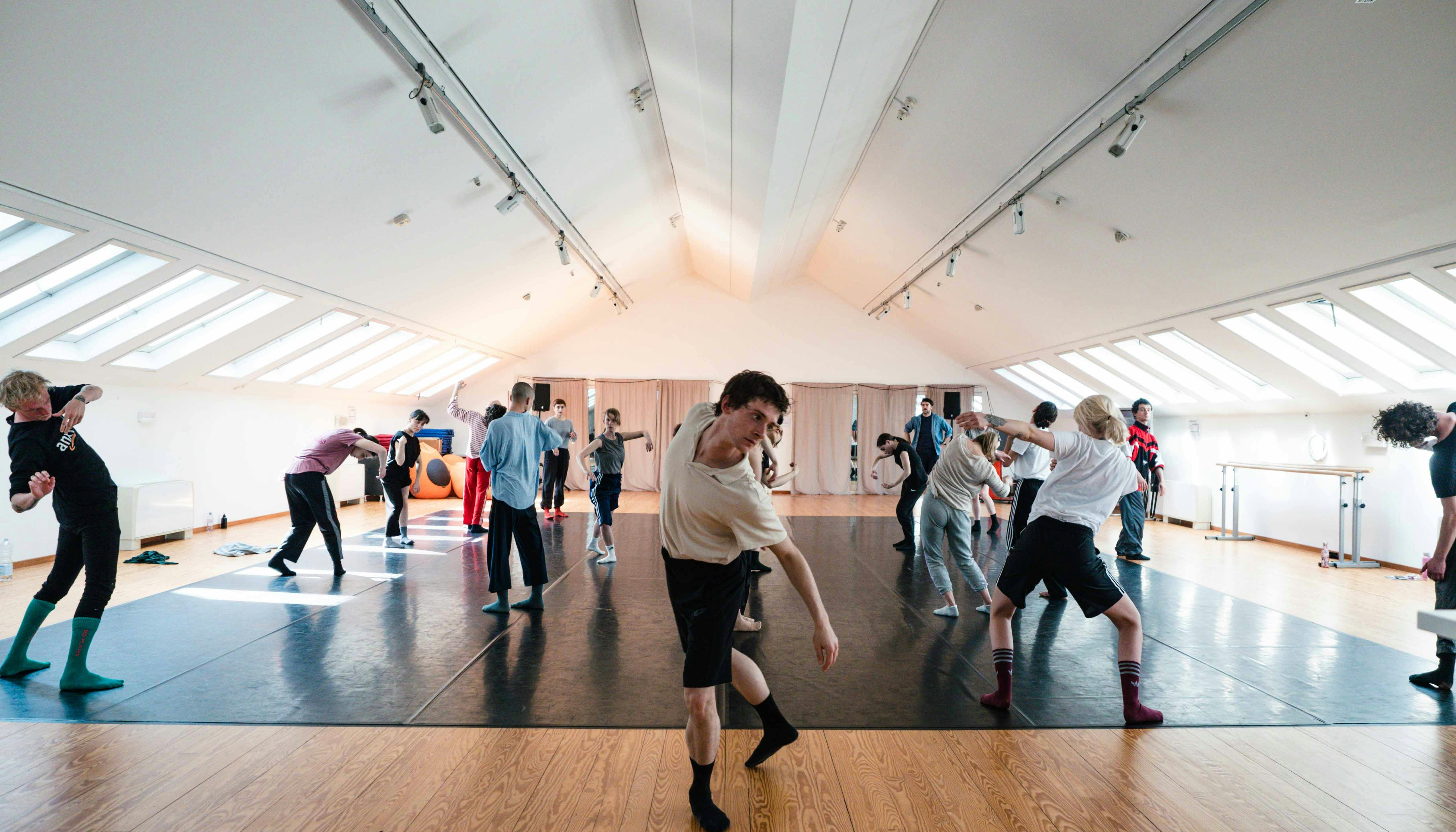 Un gruppo di danzatori in vestiti comodi sperimentano  muovendosi nello Studio di Oriente Occidente. In primo piano un giovane danzatore con pantaloncini e calzini neri e maglietta bianca si muove con il busto in avanti