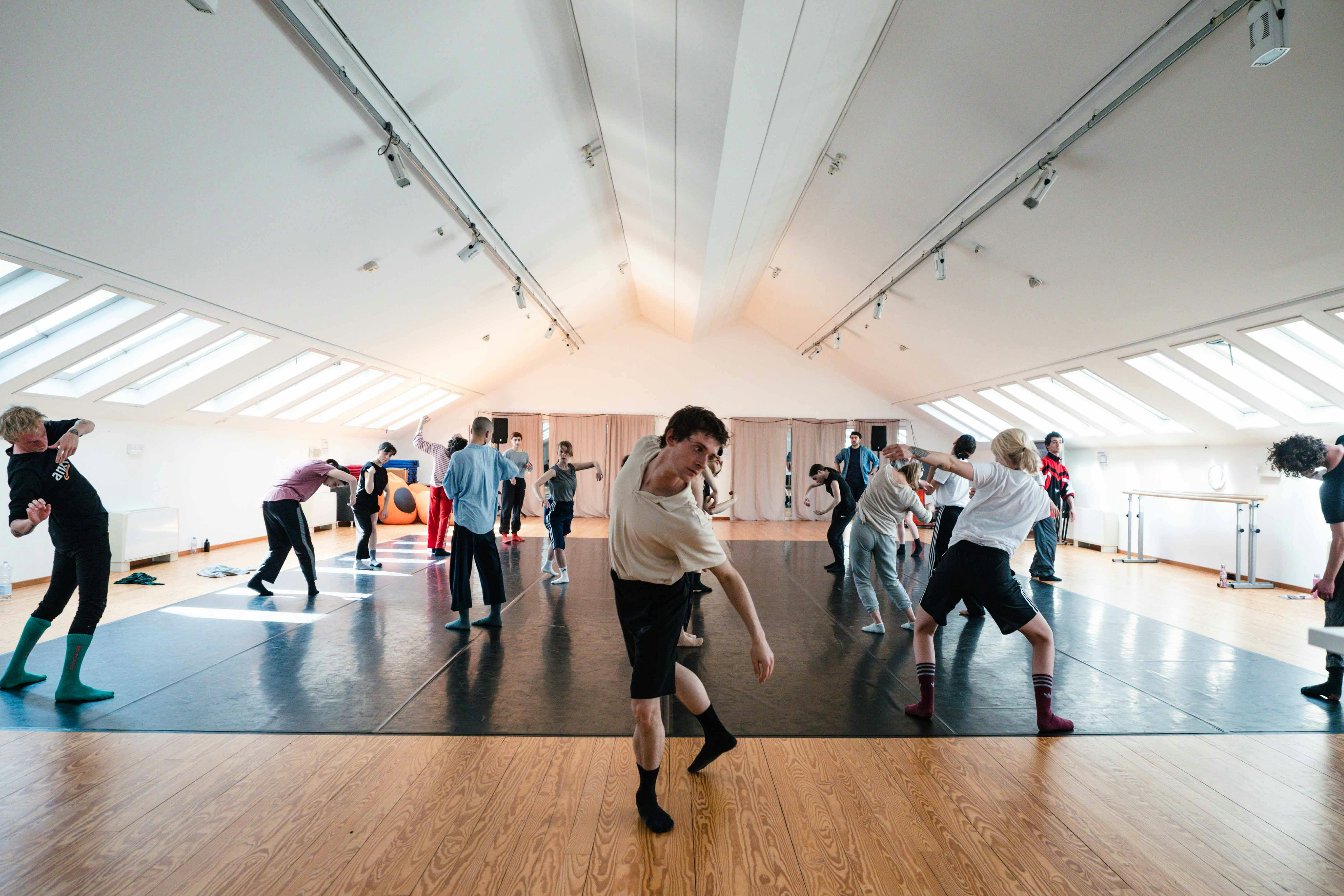 Un gruppo di danzatori in vestiti comodi sperimentano  muovendosi nello Studio di Oriente Occidente. In primo piano un giovane danzatore con pantaloncini e calzini neri e maglietta bianca si muove con il busto in avanti