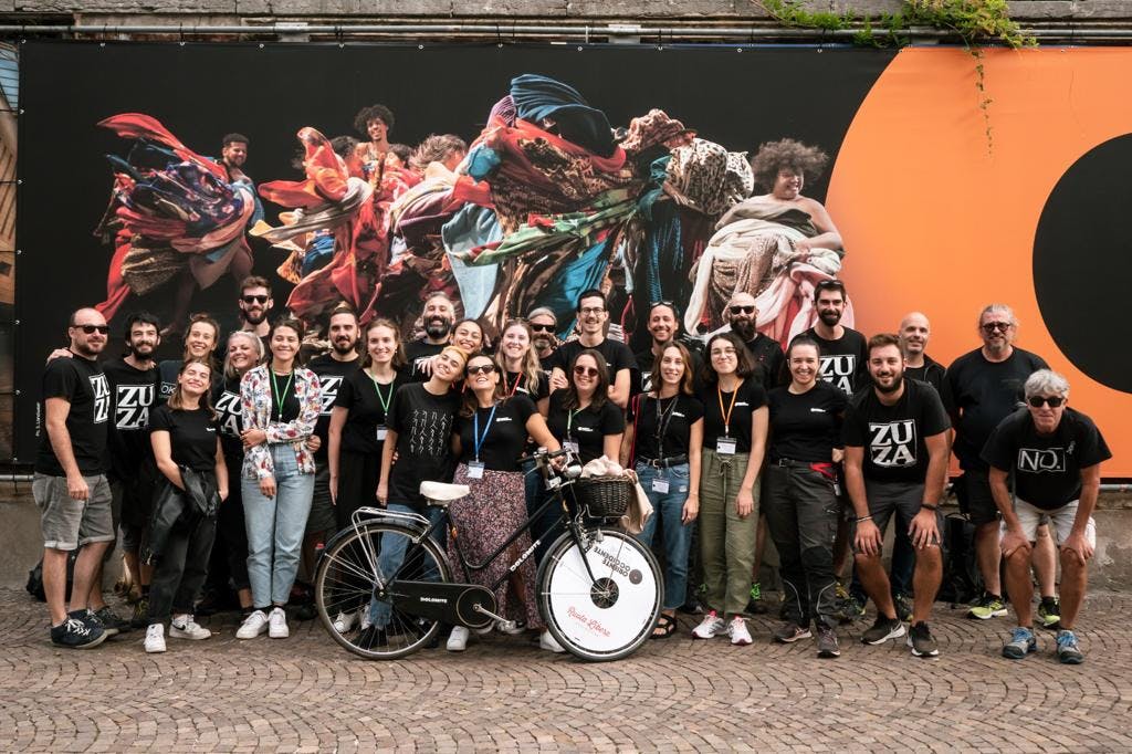 Un gruppo numeroso di collaboratori e collaboratrici dell'edizione 2022 di Oriente Occidente Dance Festival sono abbracciati sotto il poster che comunica l'evento. Indossano magliette nere e una persona in prima fila tiene la bicicletta sponsorizzata Oriente Occidente