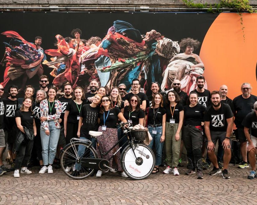 Un gruppo numeroso di collaboratori e collaboratrici dell'edizione 2022 di Oriente Occidente Dance Festival sono abbracciati sotto il poster che comunica l'evento. Indossano magliette nere e una persona in prima fila tiene la bicicletta sponsorizzata Oriente Occidente