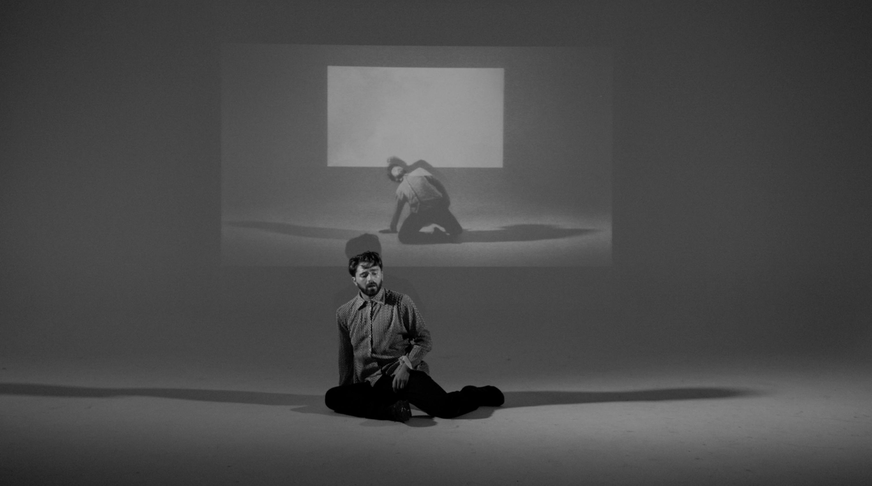 Simone Donati in una fotografia in bianco e nero indossa una camicia ed è seduto per terra, un proiettore mostra la stessa scena alle sue spalle