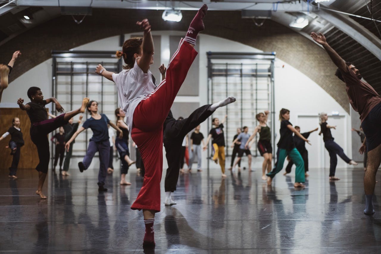 Una danzatrice con i pantaloni rossi in primo piano alza la gamba destra in secondo piano numerosi performer eseguono movimenti