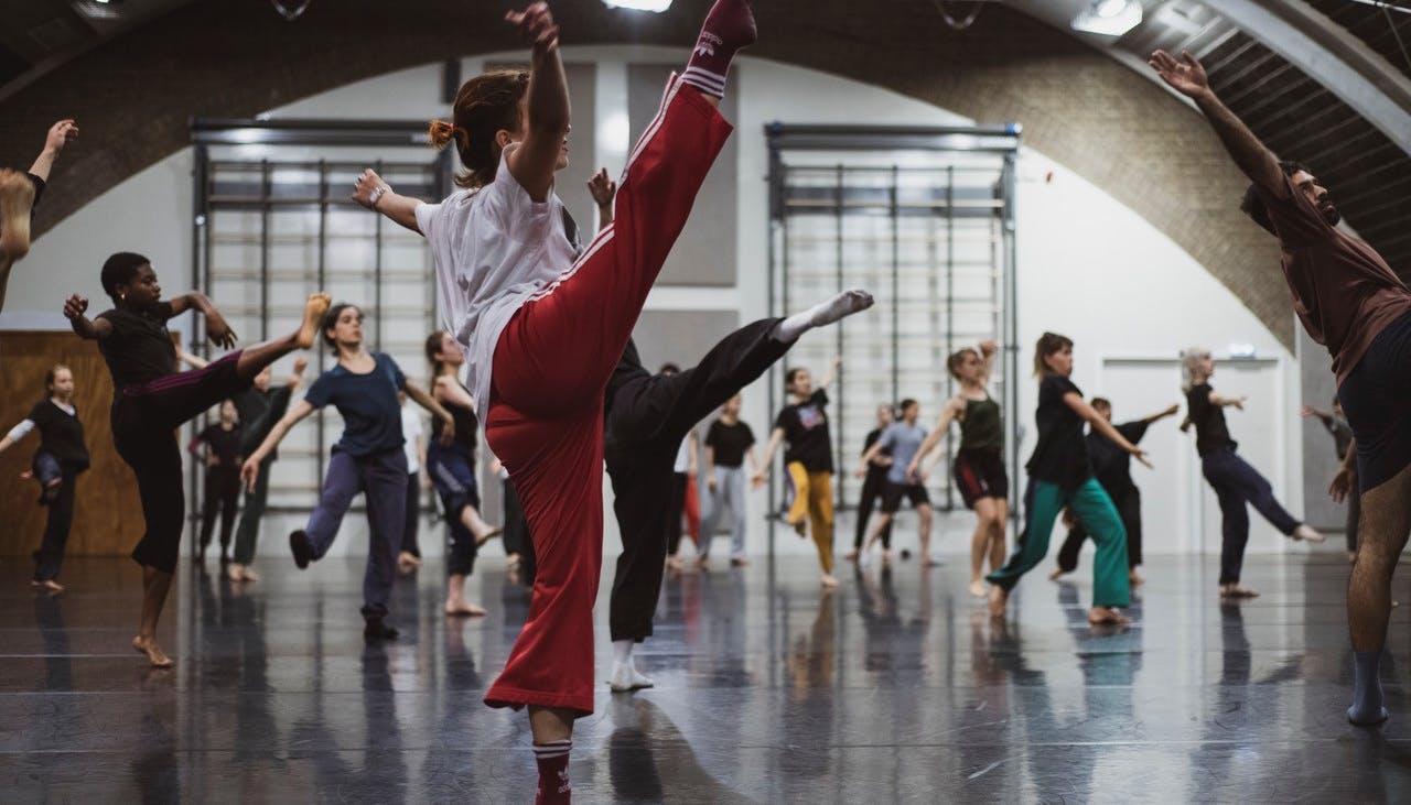 Una danzatrice con i pantaloni rossi in primo piano alza la gamba destra in secondo piano numerosi performer eseguono movimenti