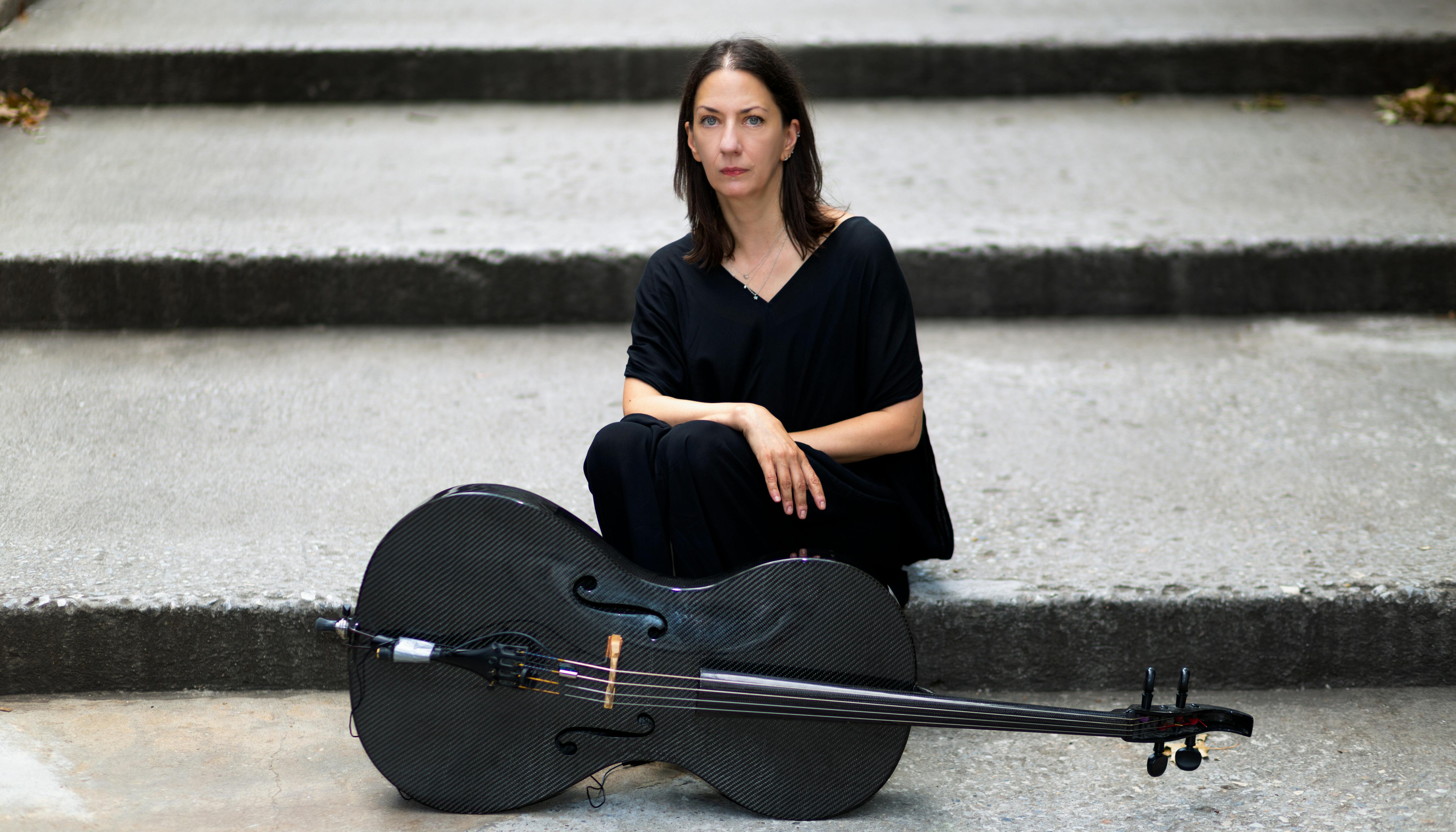 Julia Kent sitting on a staircase dressed in black with a cello in front of her