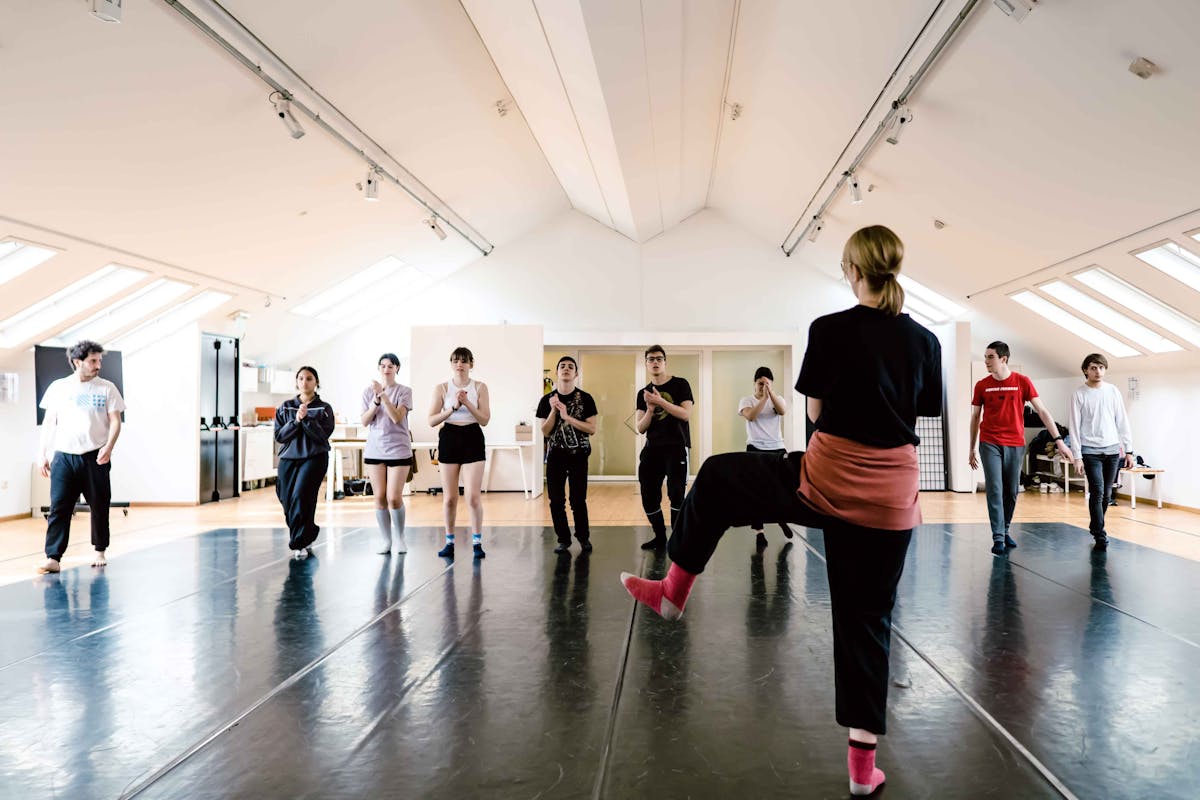 Boys and girls in the room during the workshop rehearsing a scene: in the foreground a girl in the back and the others in the background in front of her.