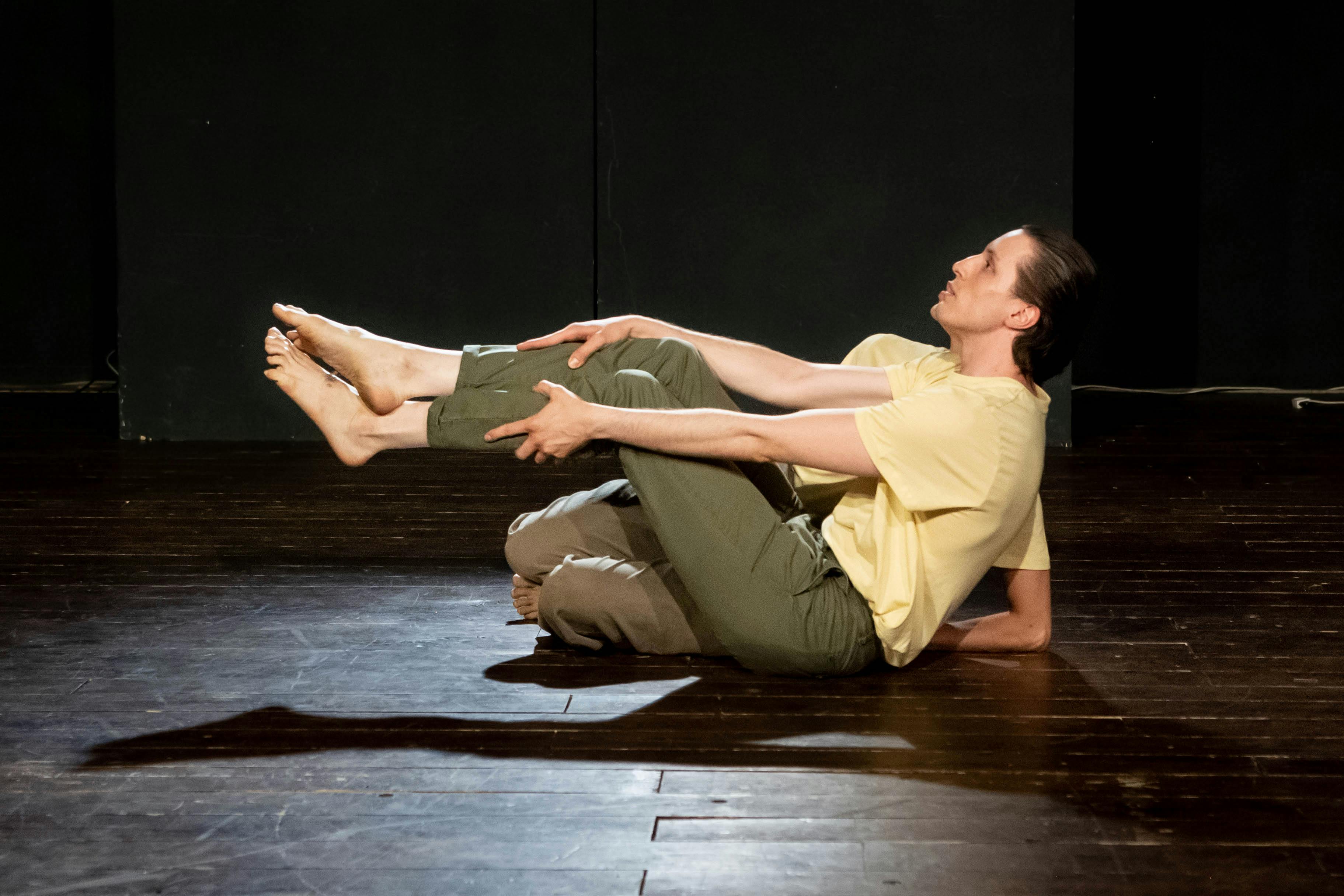 Two performers on stage wearing yellow jersey and dark green pants, one o in the foreground sitting with raised legs the second is hidden by the figure of the dancer