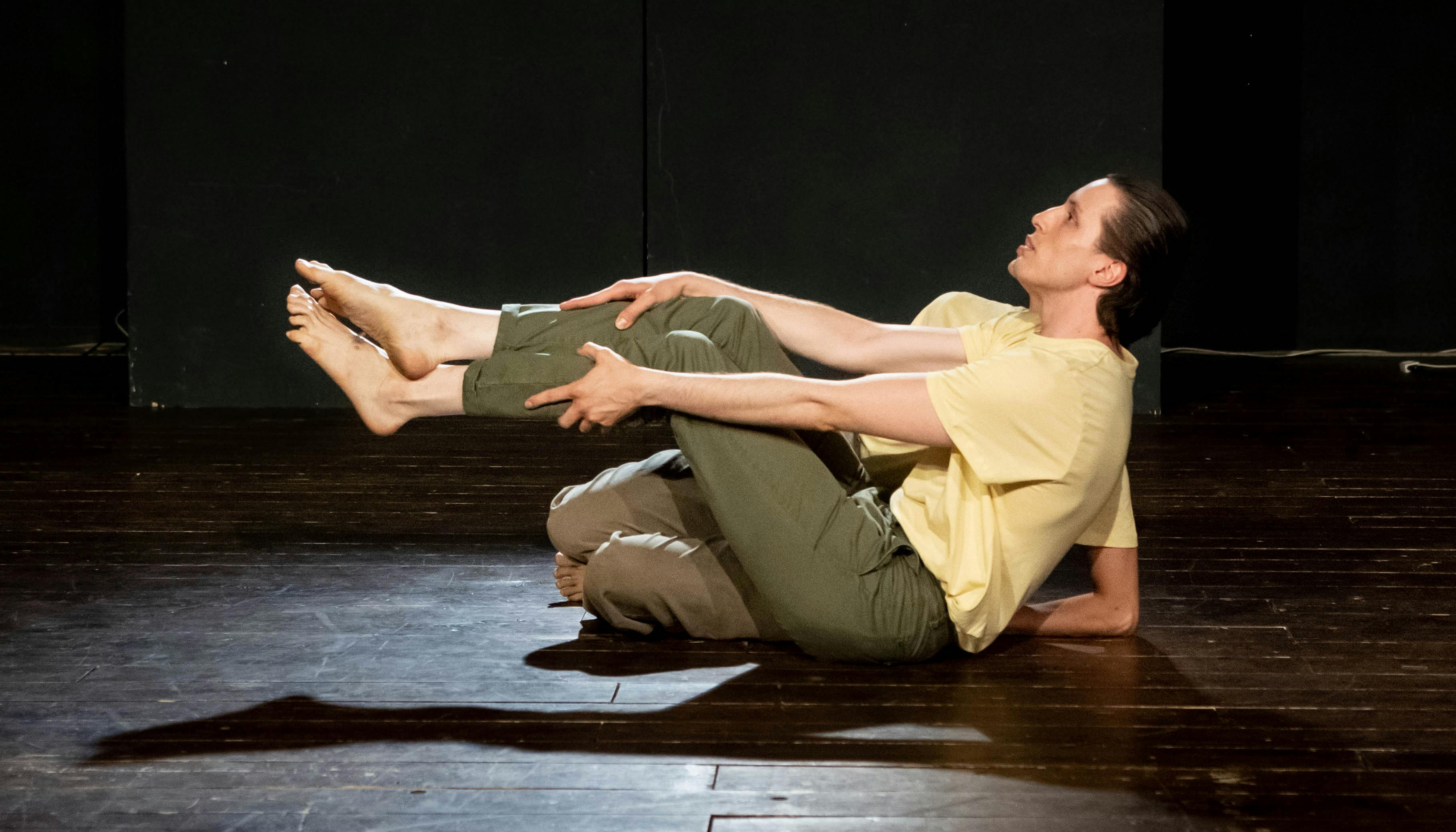 Due performer in scena con maglia gialla e pantaloni verde scuro, un o in primo piano seduto con gambe sollevate la seconda è nascosta dalla figura del danzatore