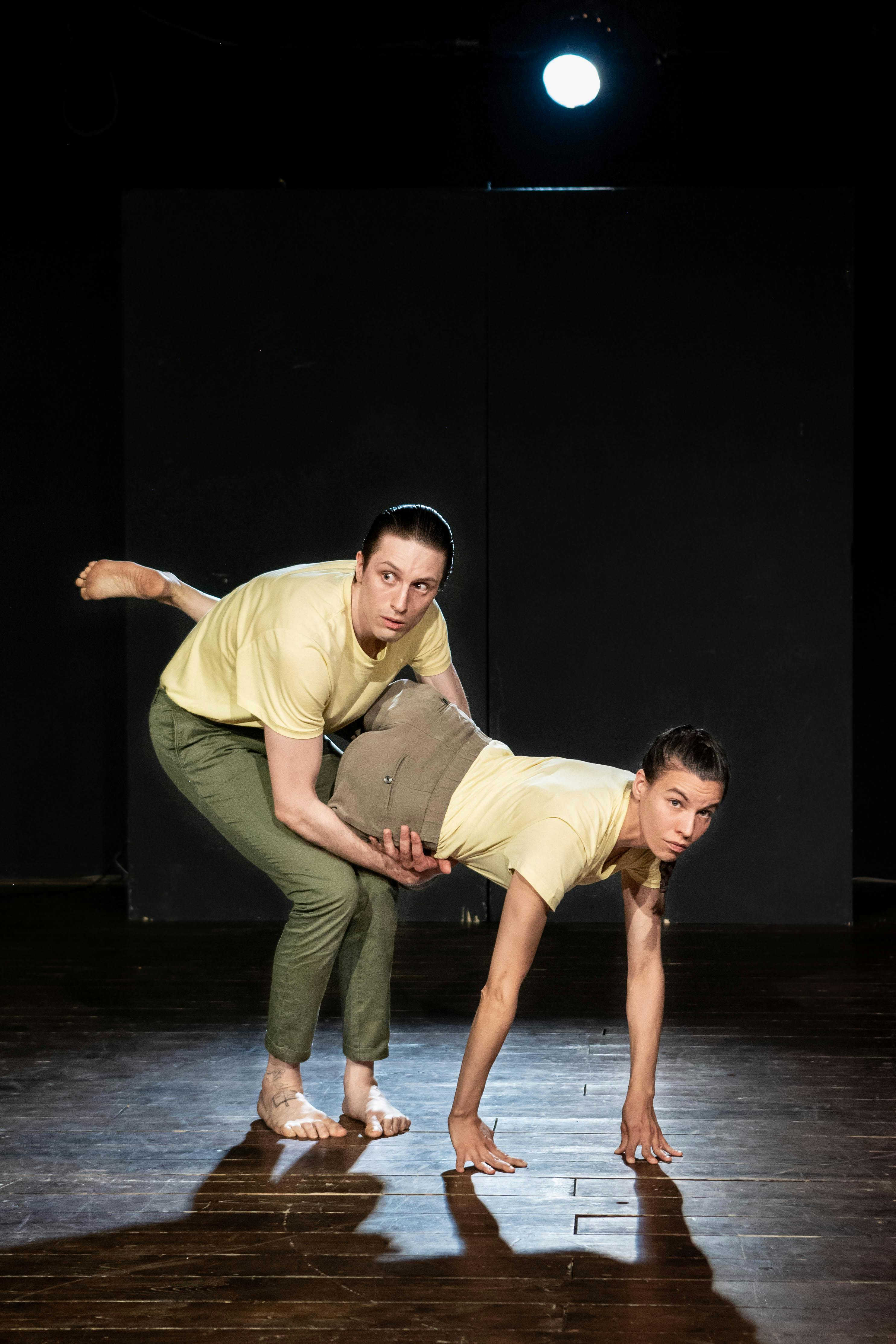 A dancer supports the pelvis of a female dancer who leans on the floor with her hands