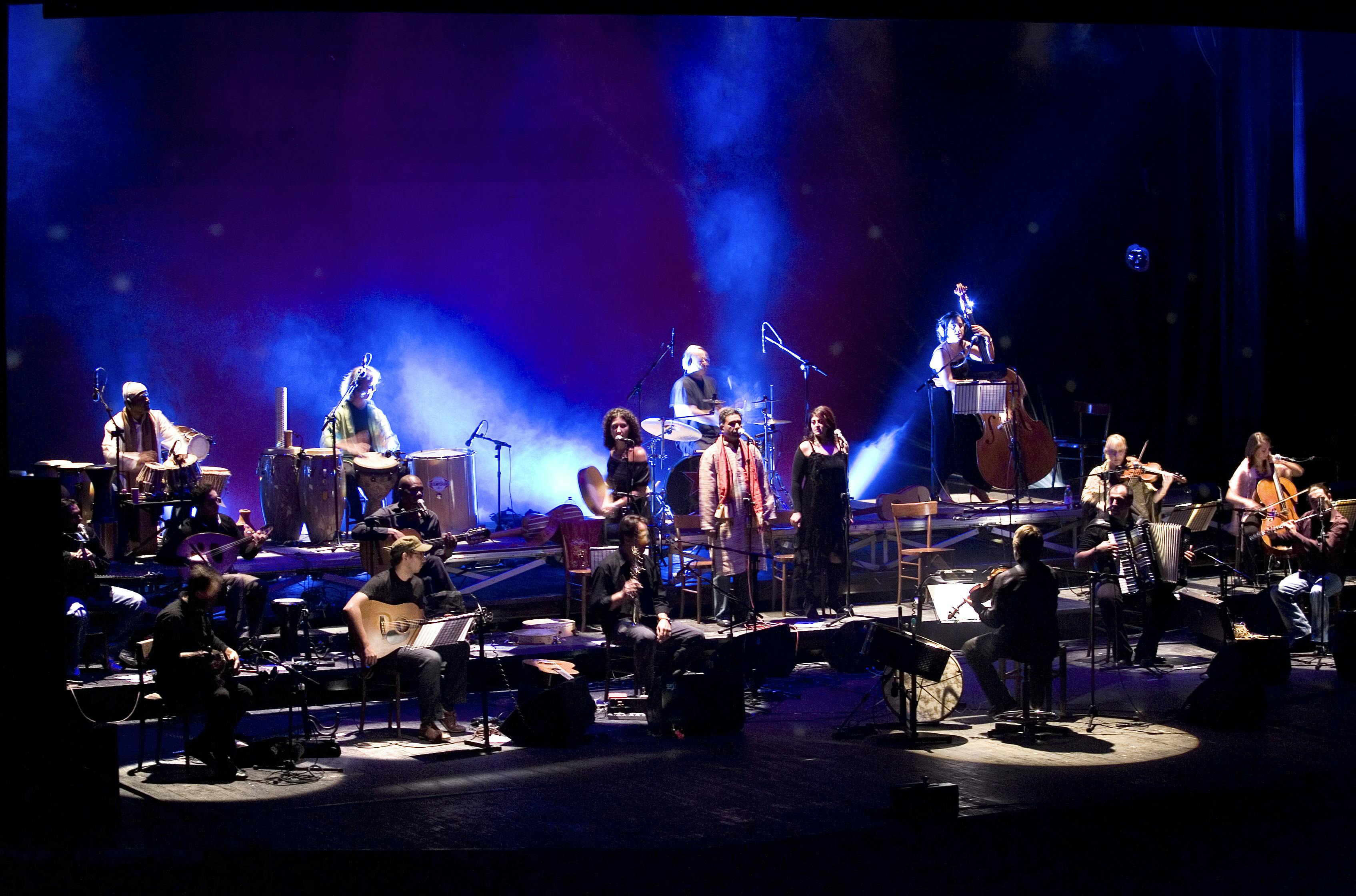 Un ampio gruppo di musicisti si esibiscono su un palco illuminati da luci di colore blu